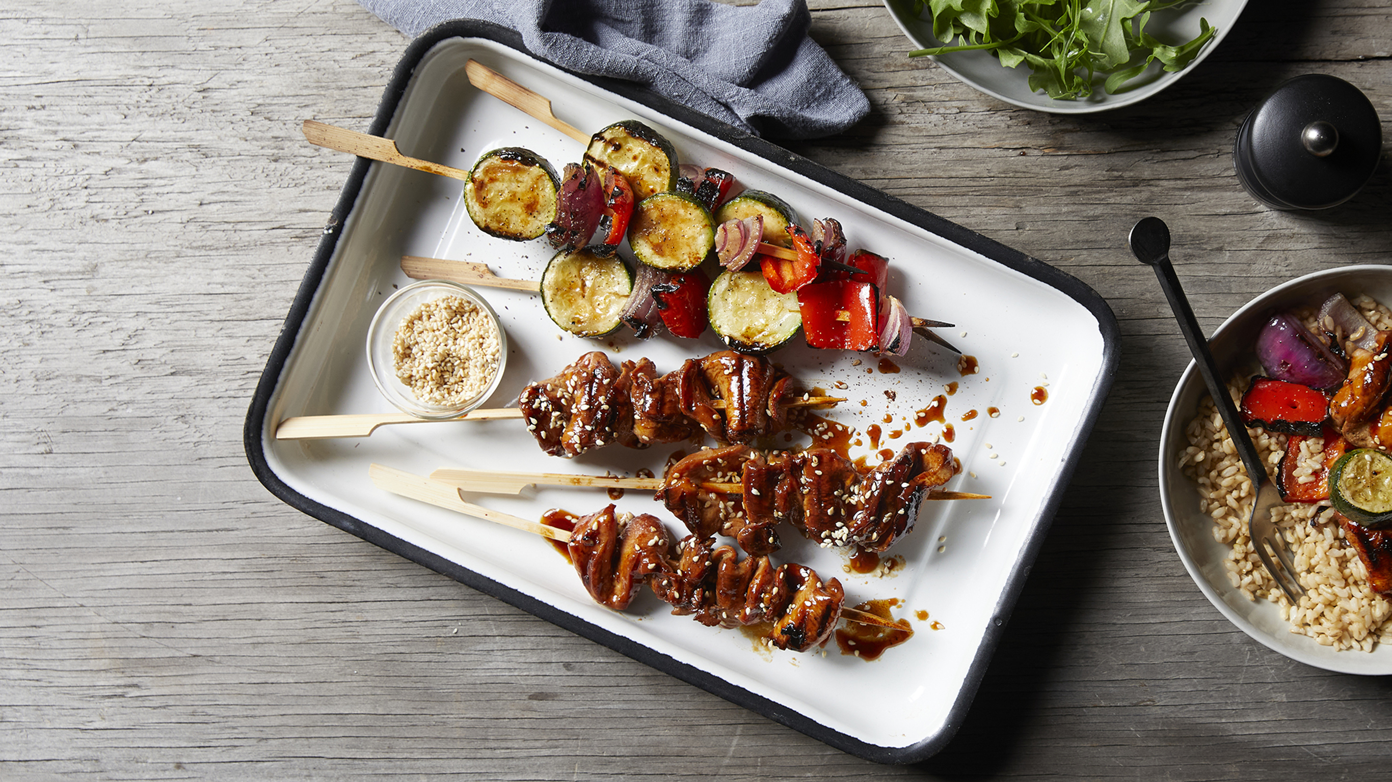 An enamel tray of teriyaki grilled skewers sits on a rustic wooden table, ready for serving