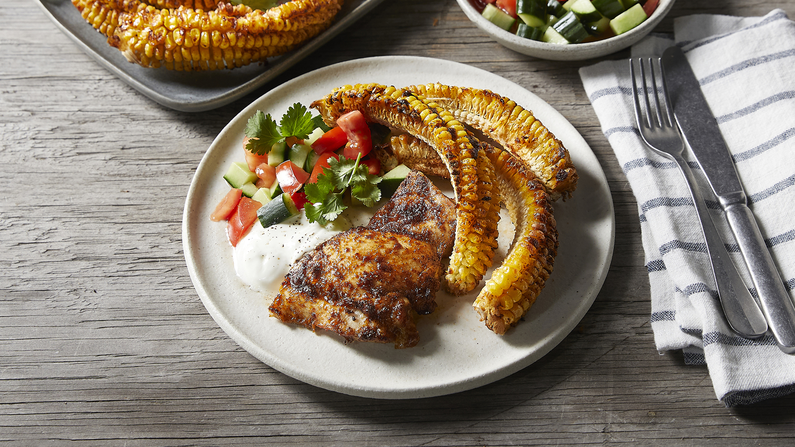 A serve of grilled chicken and spicy corn ribs sits on a rustic wooden table, with knife and fork resting on a striped napkin, ready for eating.