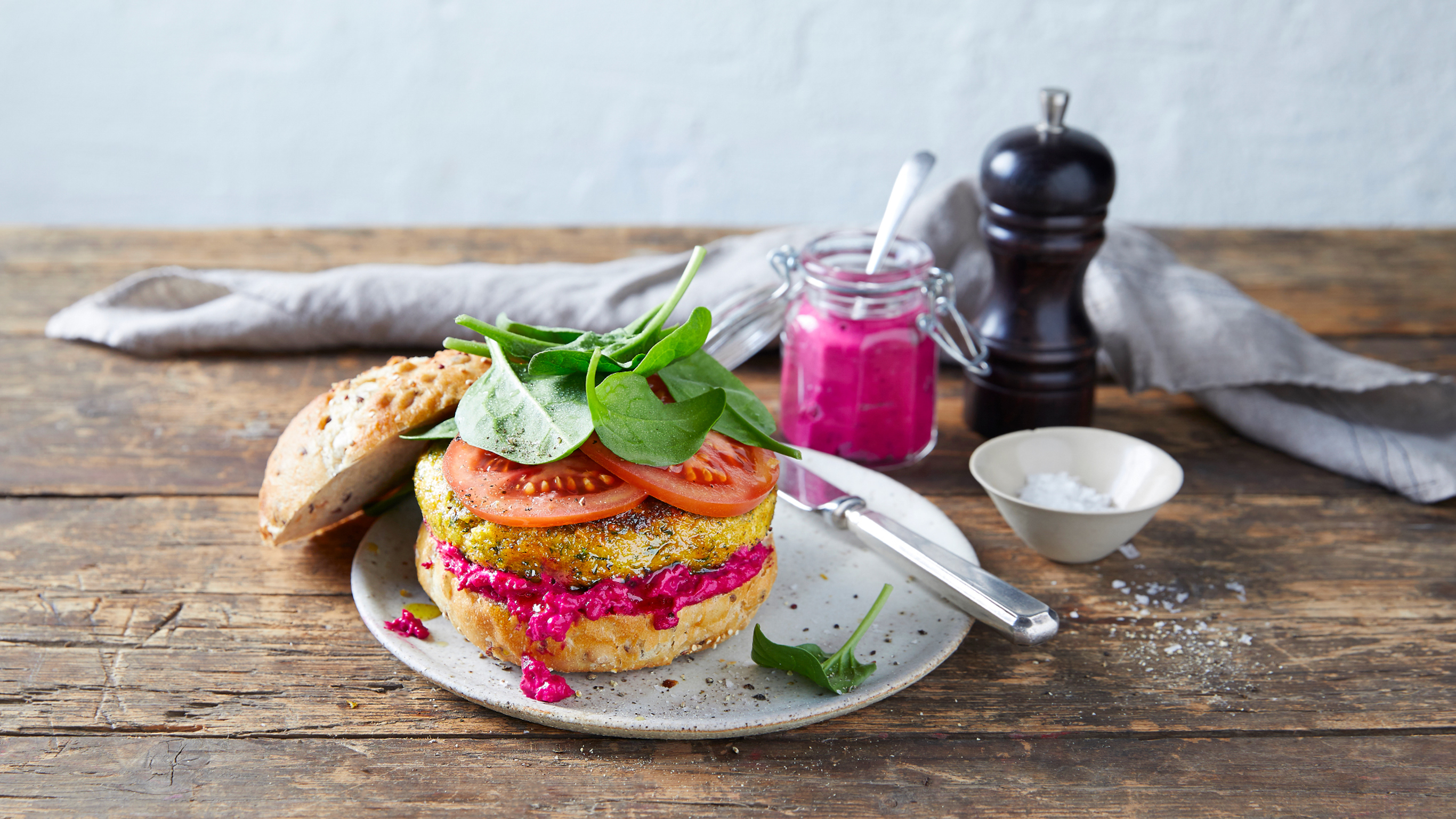A veggie burger with deep pink beetroot hummus sits on a plate, with slices of tomato and baby spinach leaves on top.