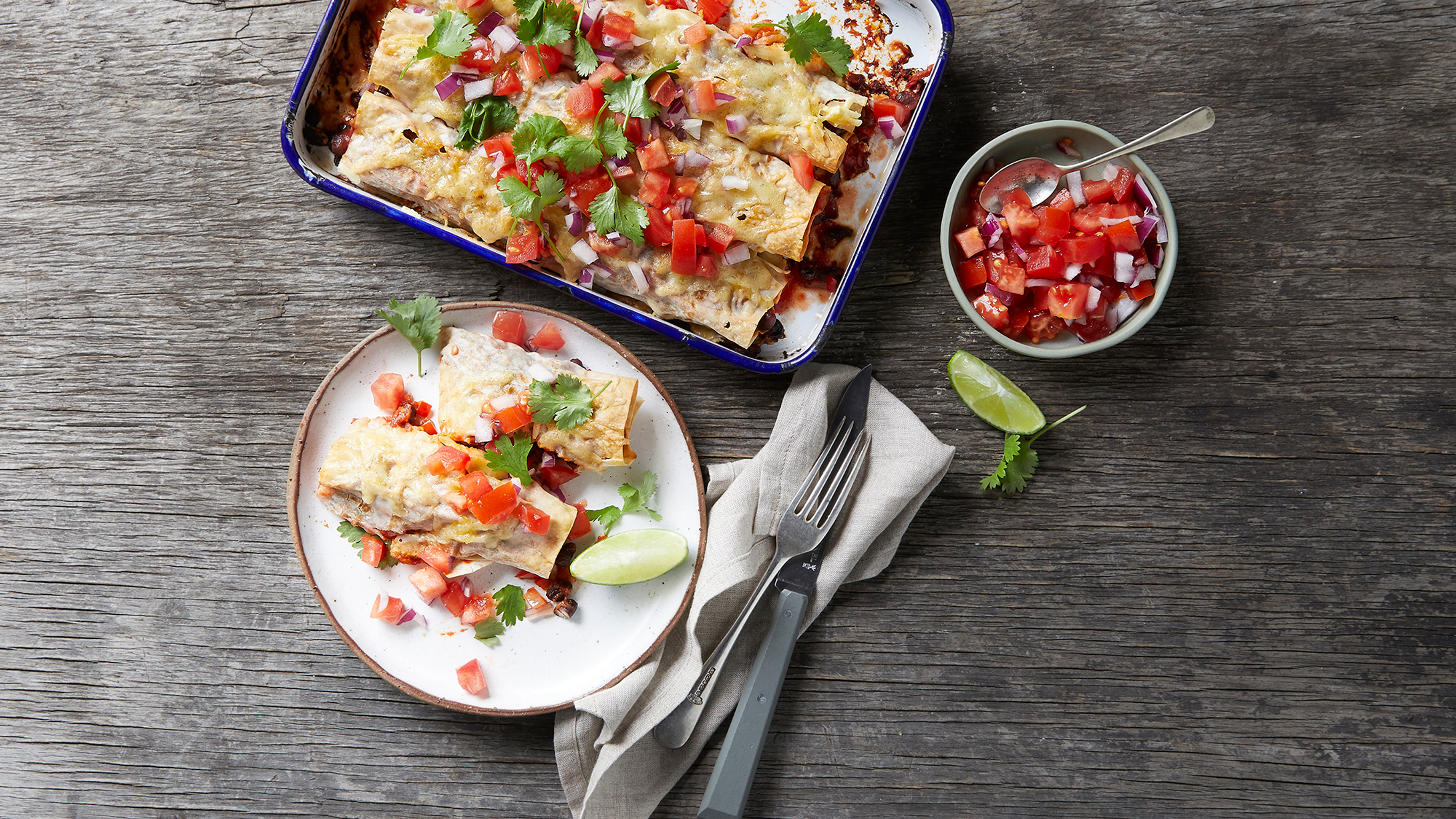A serving of Bean Enchiladas with Salsa, with knife and fork on the side,  sits beside a baking tray of enchiladas.