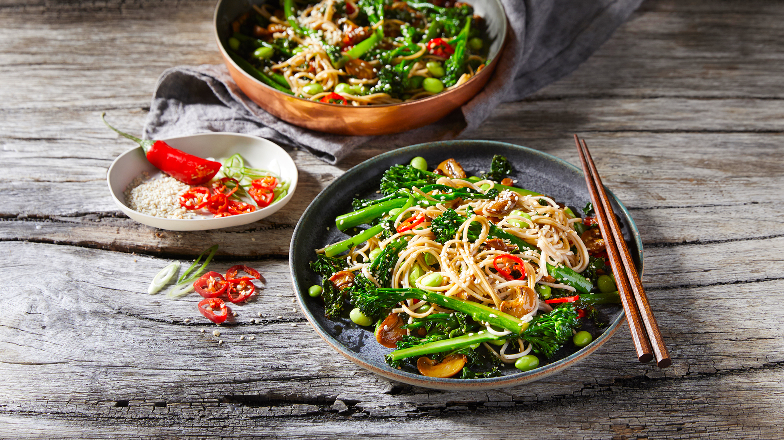 A serve of Teriyaki Soba Noodle Salad sits in front of a cooking pan of noodle salad, ready for eating.