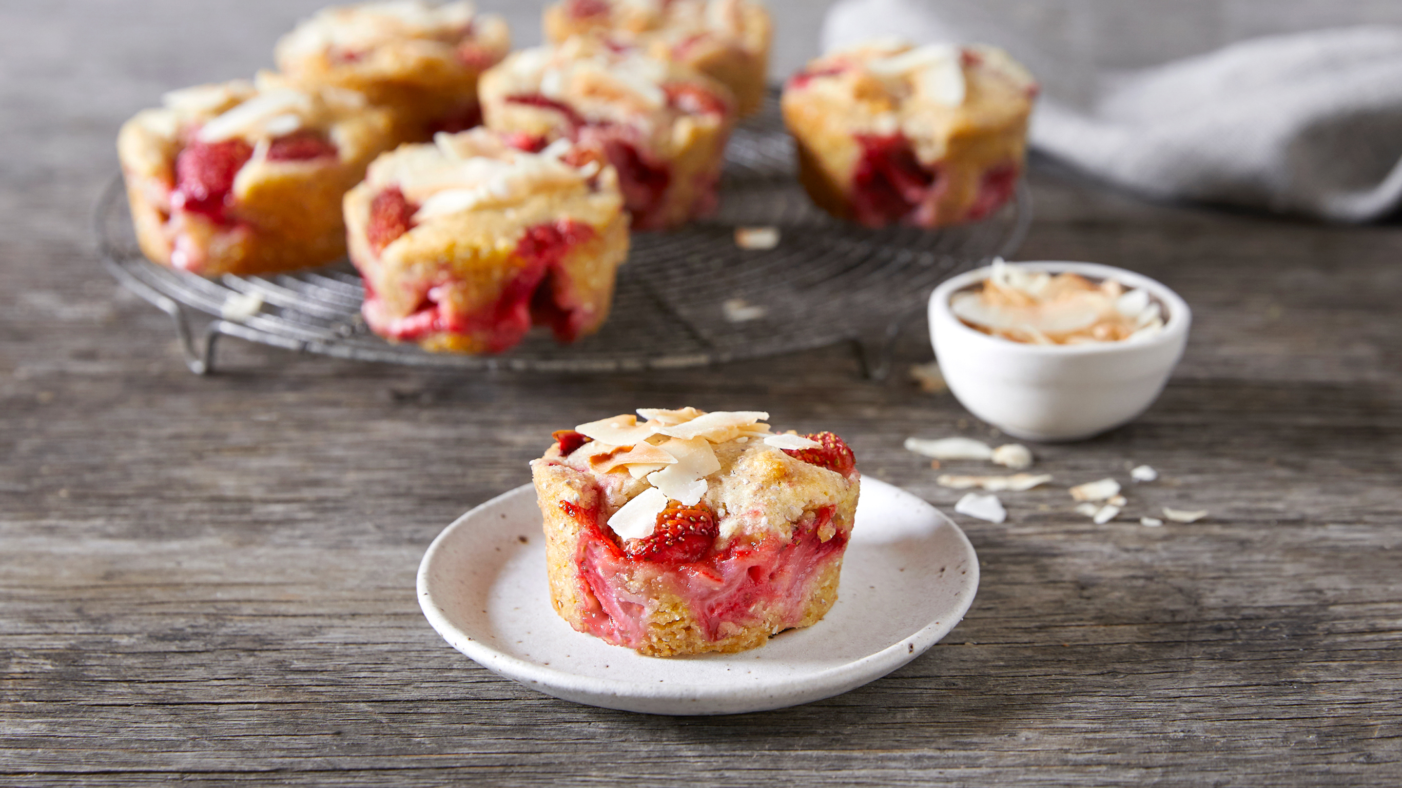A single Strawberry & Coconut Muffins sits on a small plate in front of a circular rack of muffins, with coconut flakes scattered around.