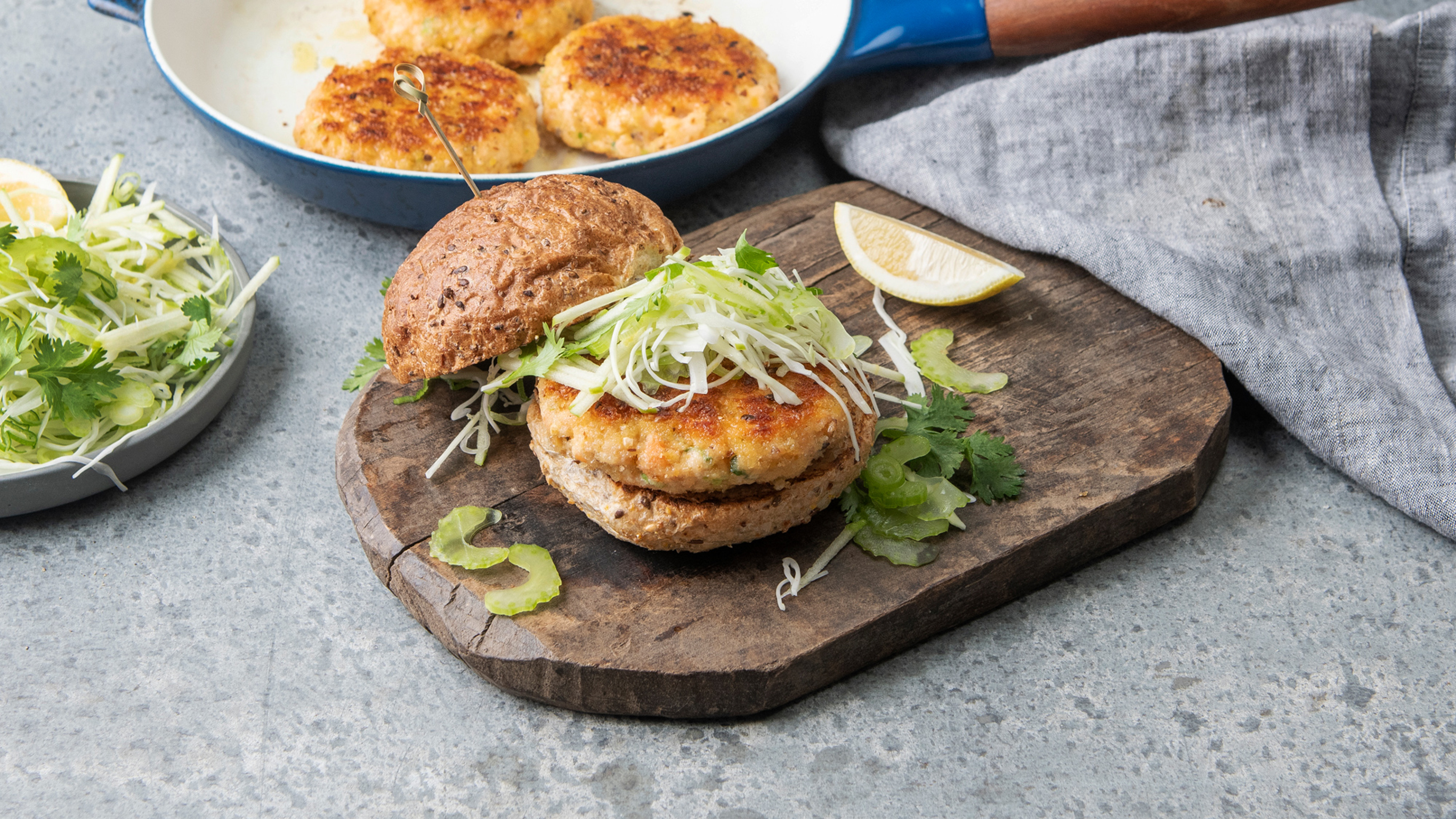 A salmon burger sits on a wooden board, with salmon burger patties in the background.