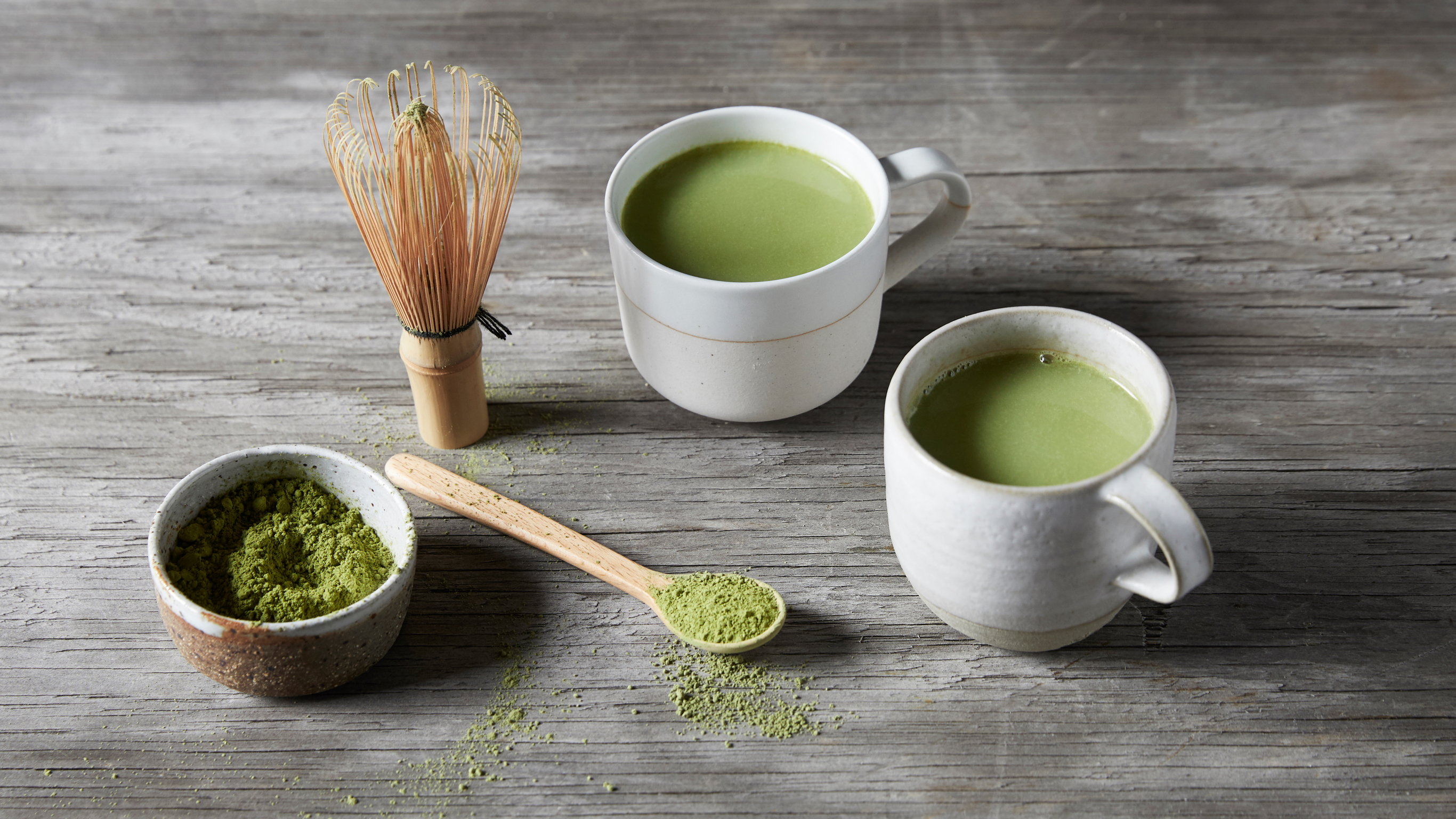 Two mugs of Vegan Matcha Latte sit on a wooden table, with a small bowl of matcha powder and a wooden spoon with matcha powder on the side.