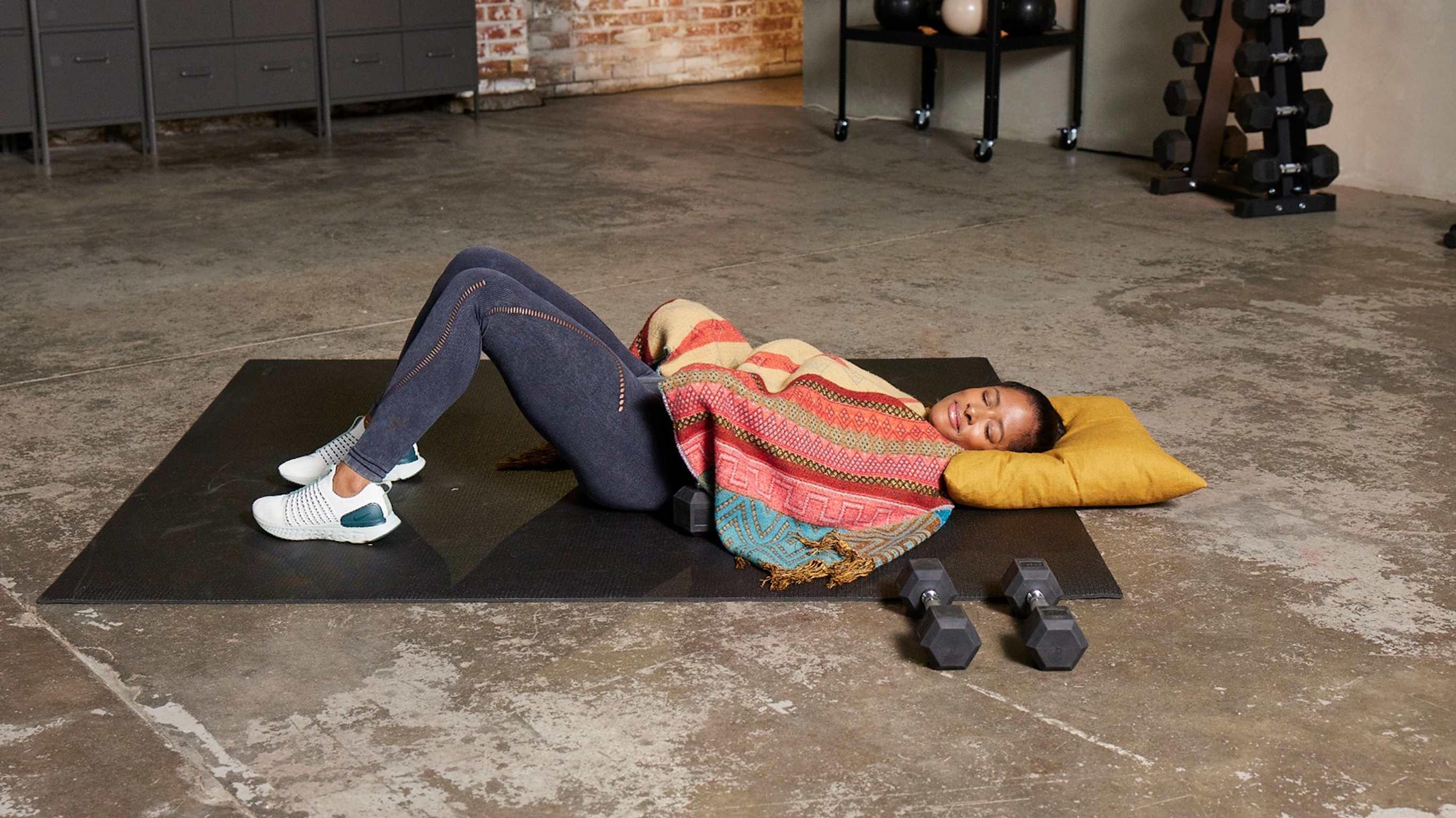 Ingrid Clay, wrapped in a colourful blanket, lies with her eyes closed on a yoga mat, head on a pillow and a set of dumbbells to one side.