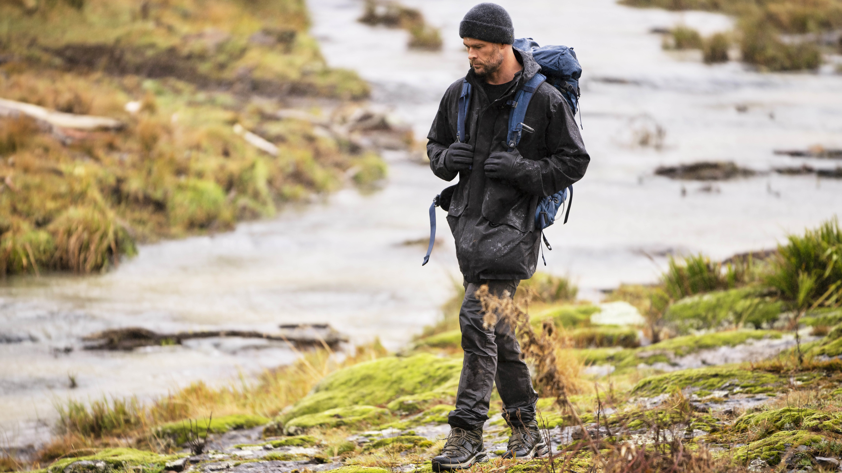 Centr founder Chris Hemsworth, carrying a backpack and wearing winter clothing, hikes along a trail next to a stream.