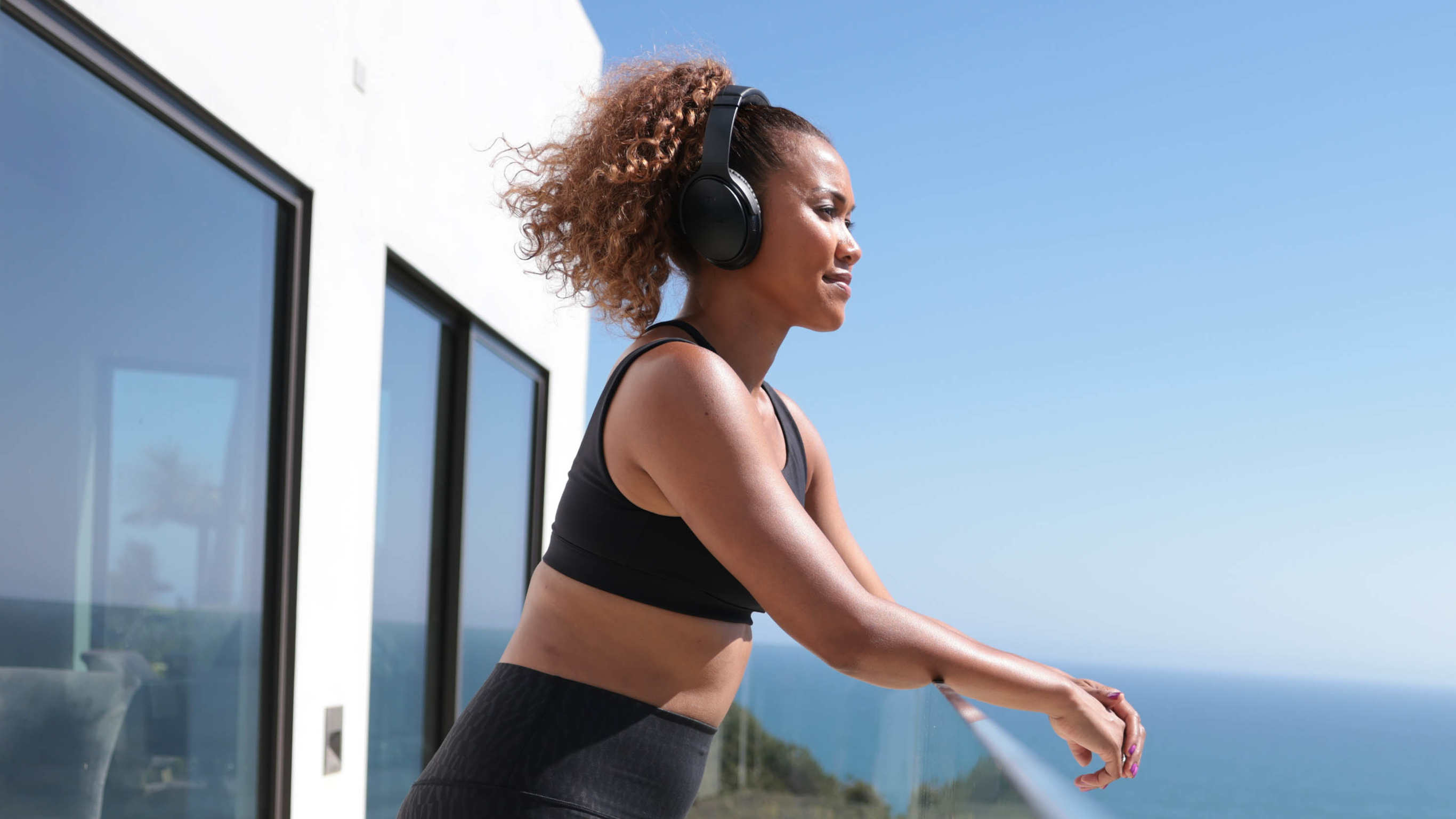 Centr beginner fitness trainer Maricris Lapaix, wearing heaphones, leans on a balcony railing and looks out towards the ocean.