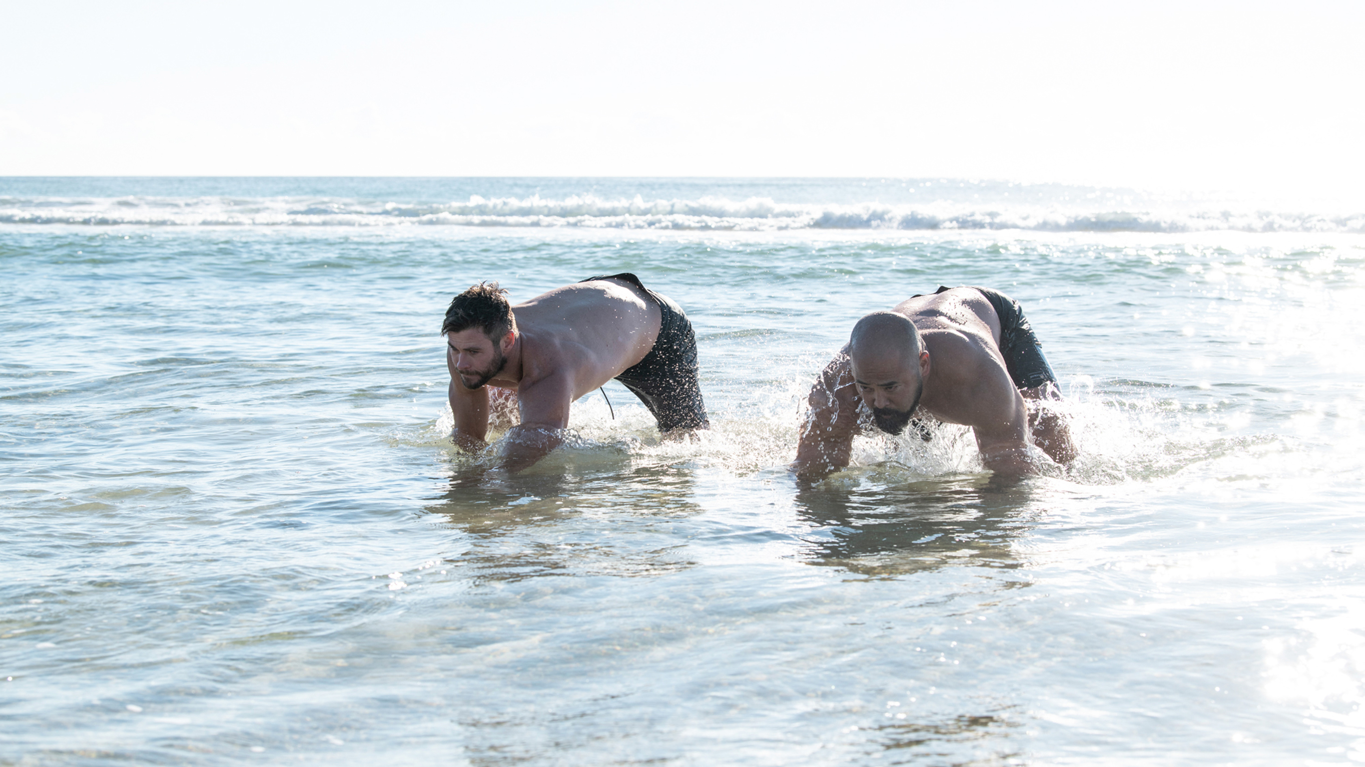 Chris Hemsworth and Da Rulk doing bear crawls in the water on an ocean shore.