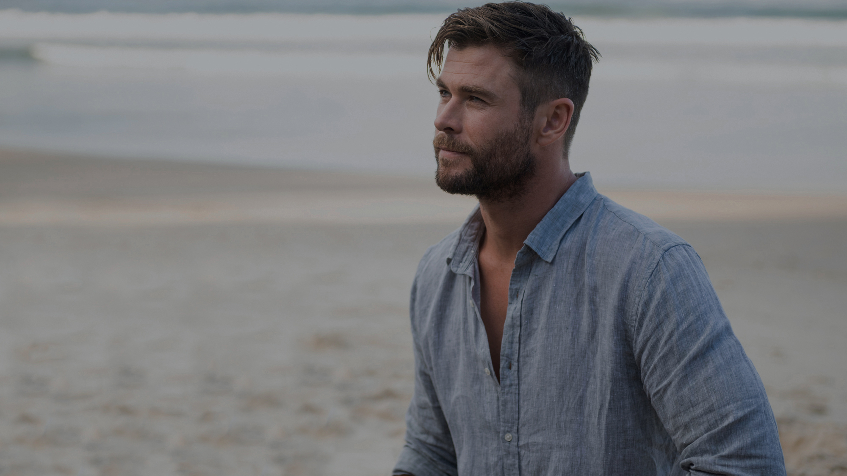 Chris Hemsworth sits on a beach wearing a blue button up shirt.