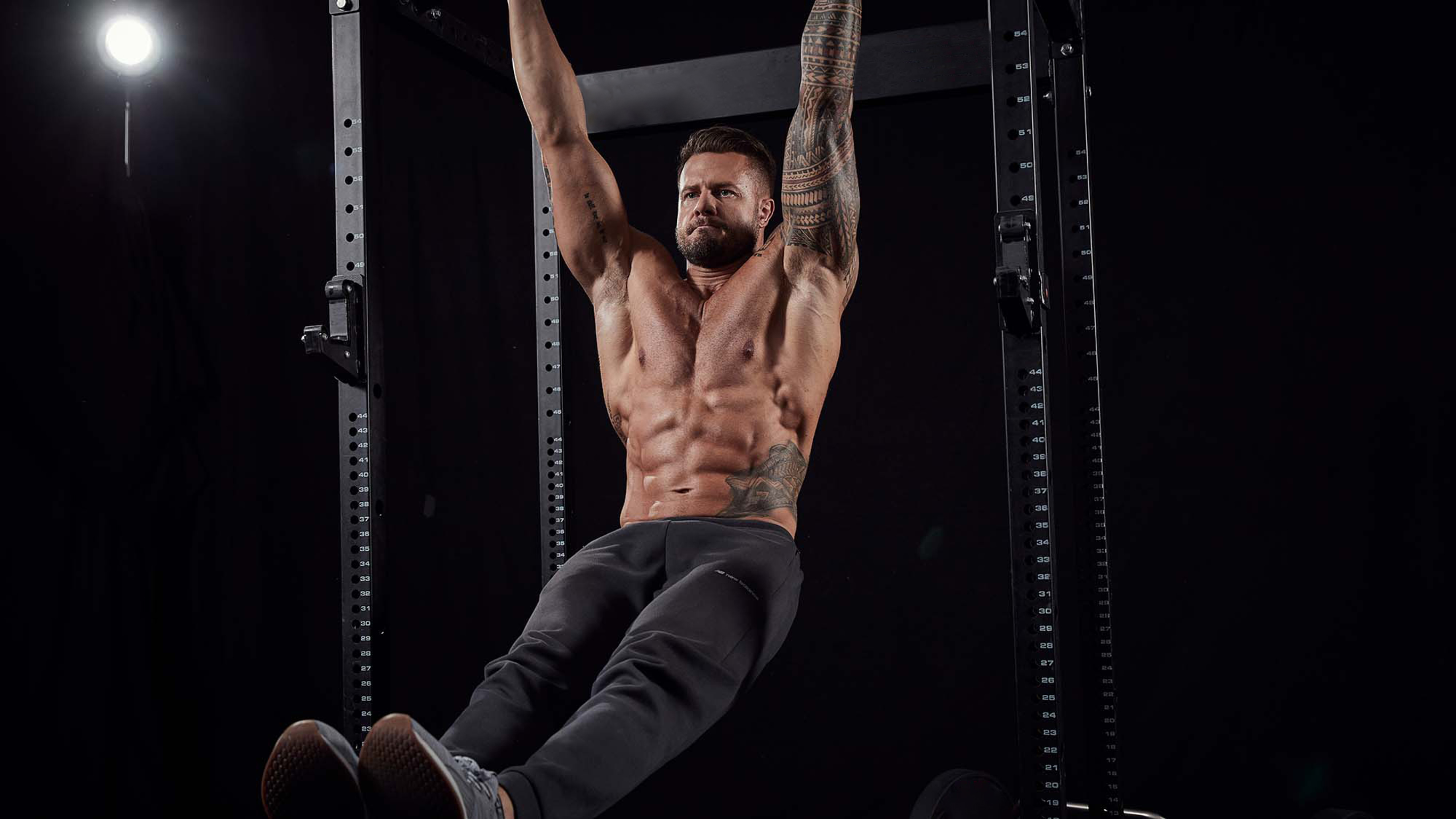 A shirtless, muscular man with visible abs hanging from a pull-up bar.