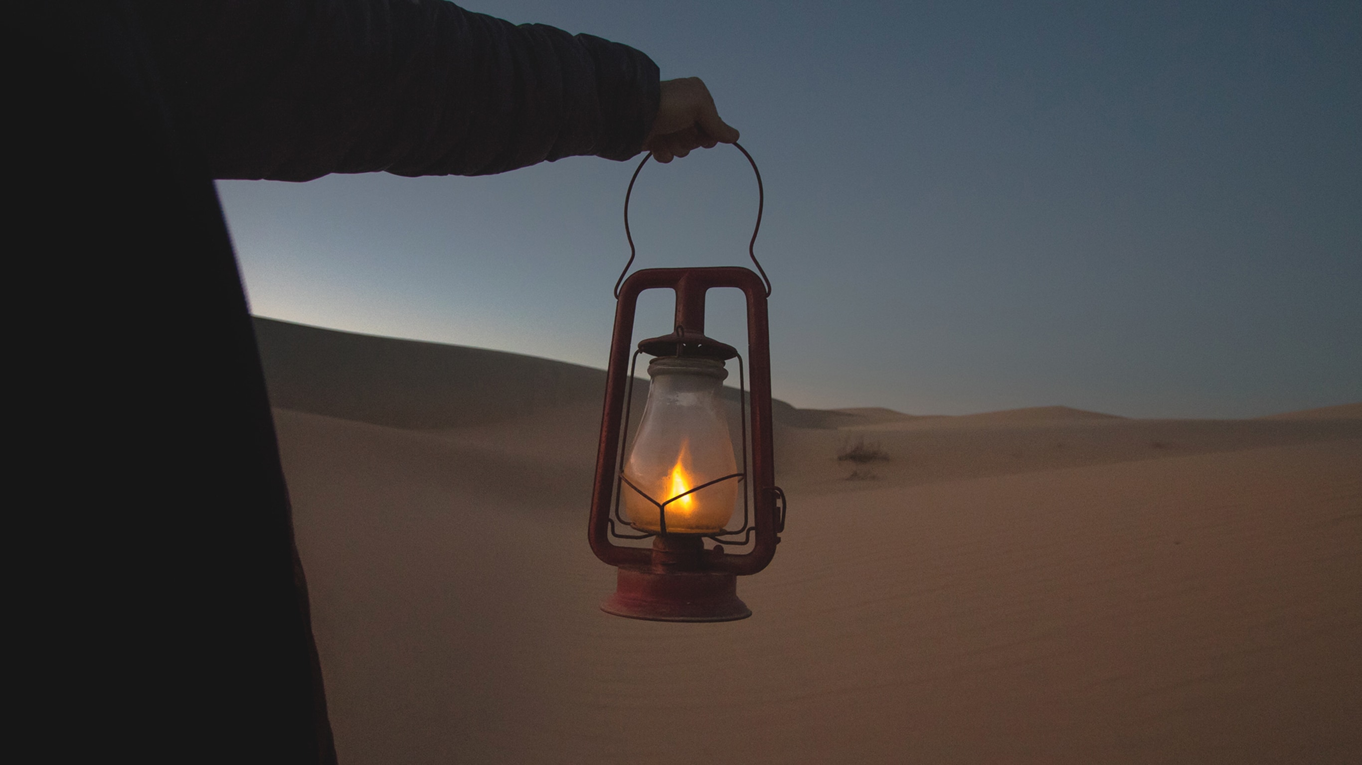 An unseen person holds a gas lantern in a sandy location.
