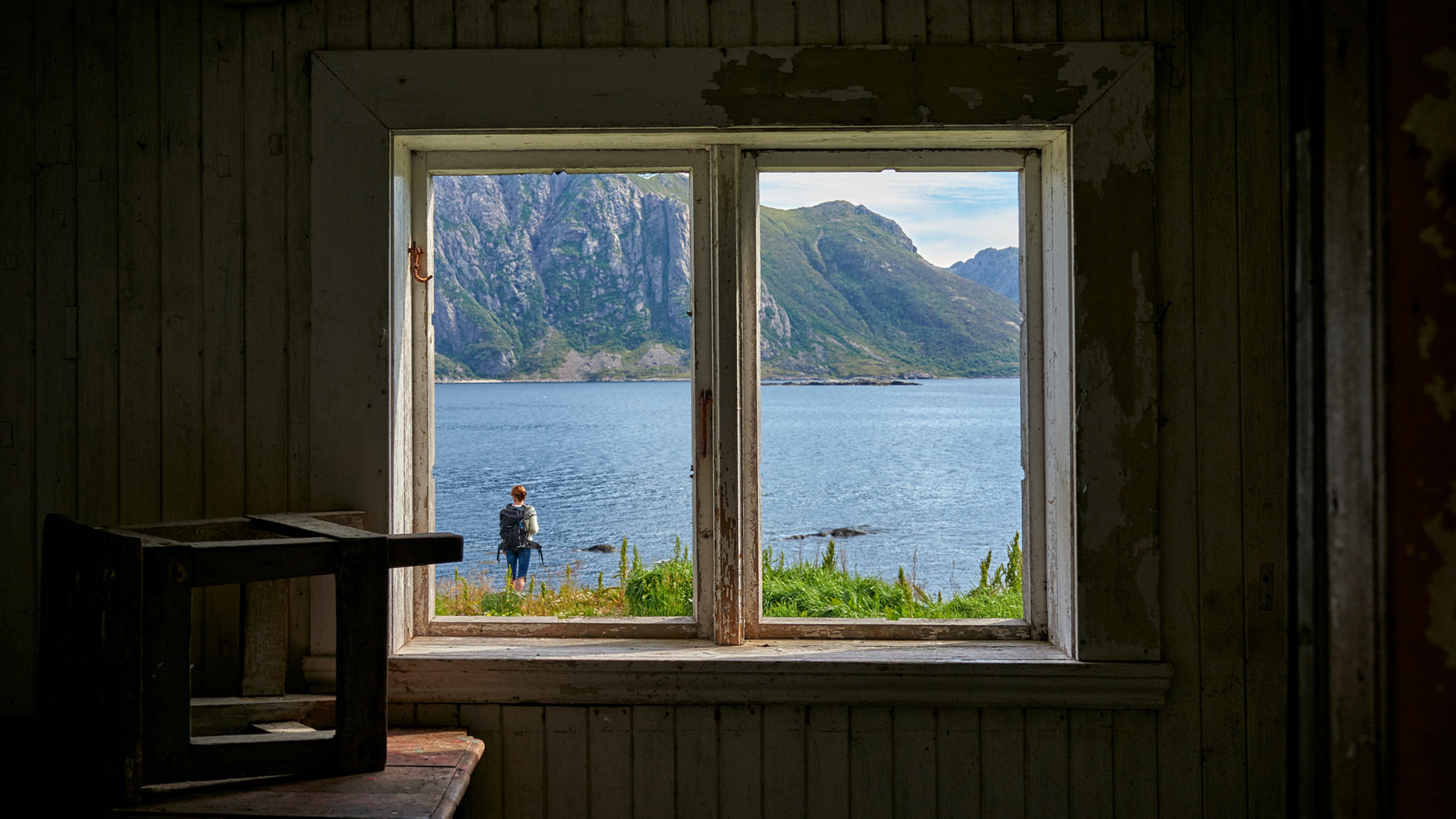 A window in an old house reveals a person with a backpack in front of a  mountain lake outside.