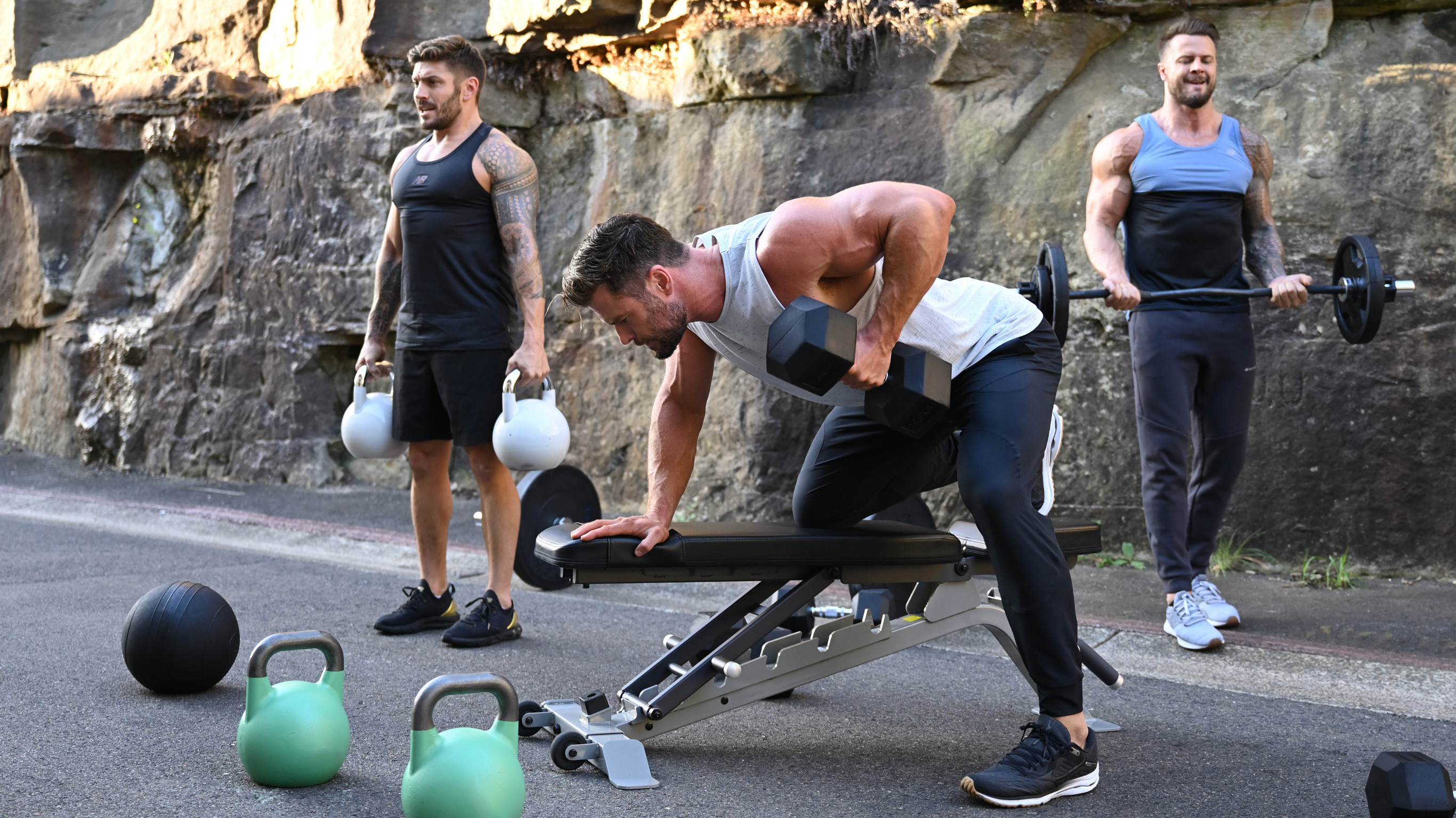 Centr trainers Luke Zocchi and Bobby Holland Hanton workout with Chris Hemsworth in a rock-faced laneway, with gym equipment around them.