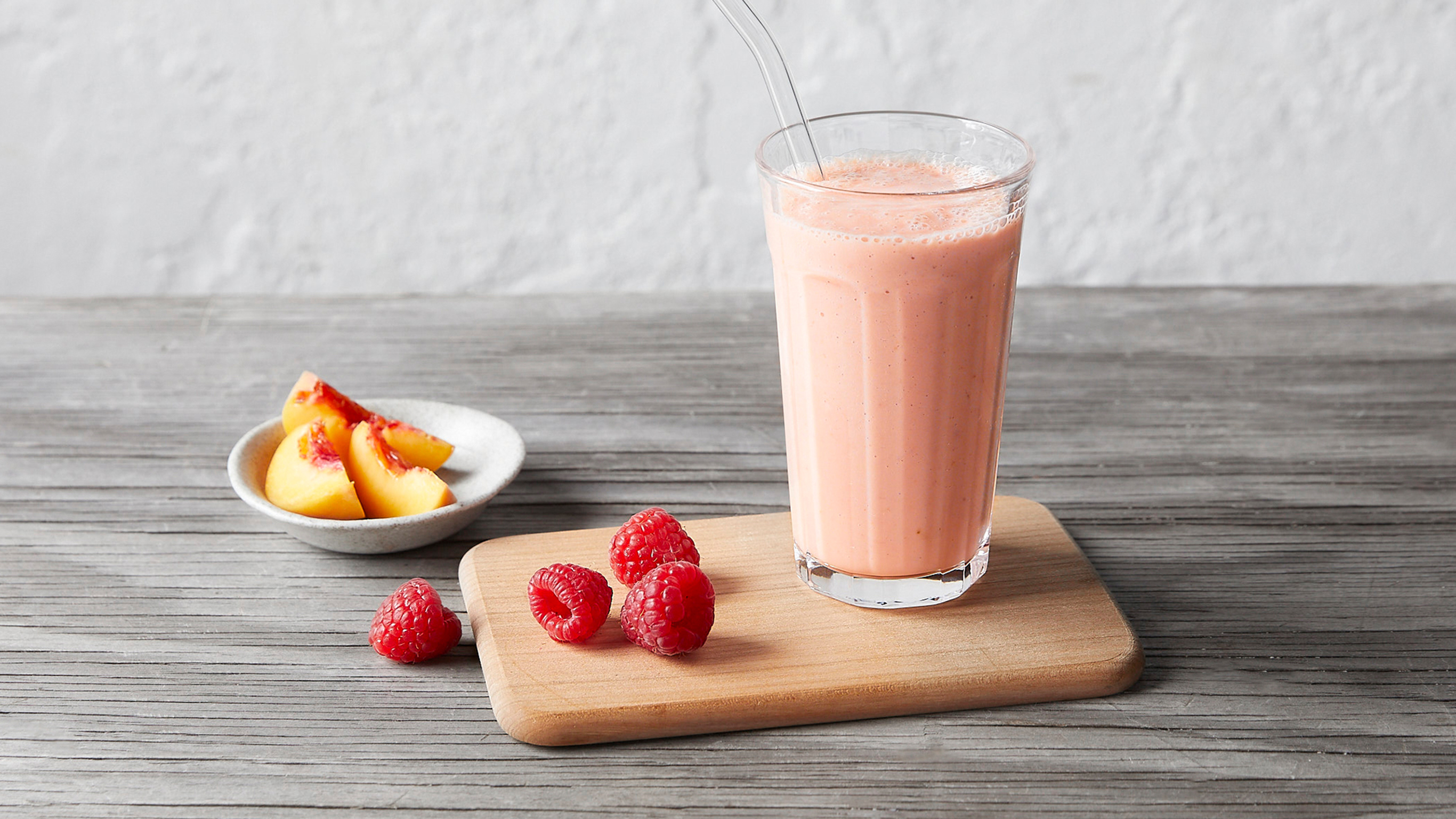 A glass of peachy pink Peach and Raspberry Smoothie stands on a wooden board, with three raspberries on the side and a small side dish of sliced peaches nearby.