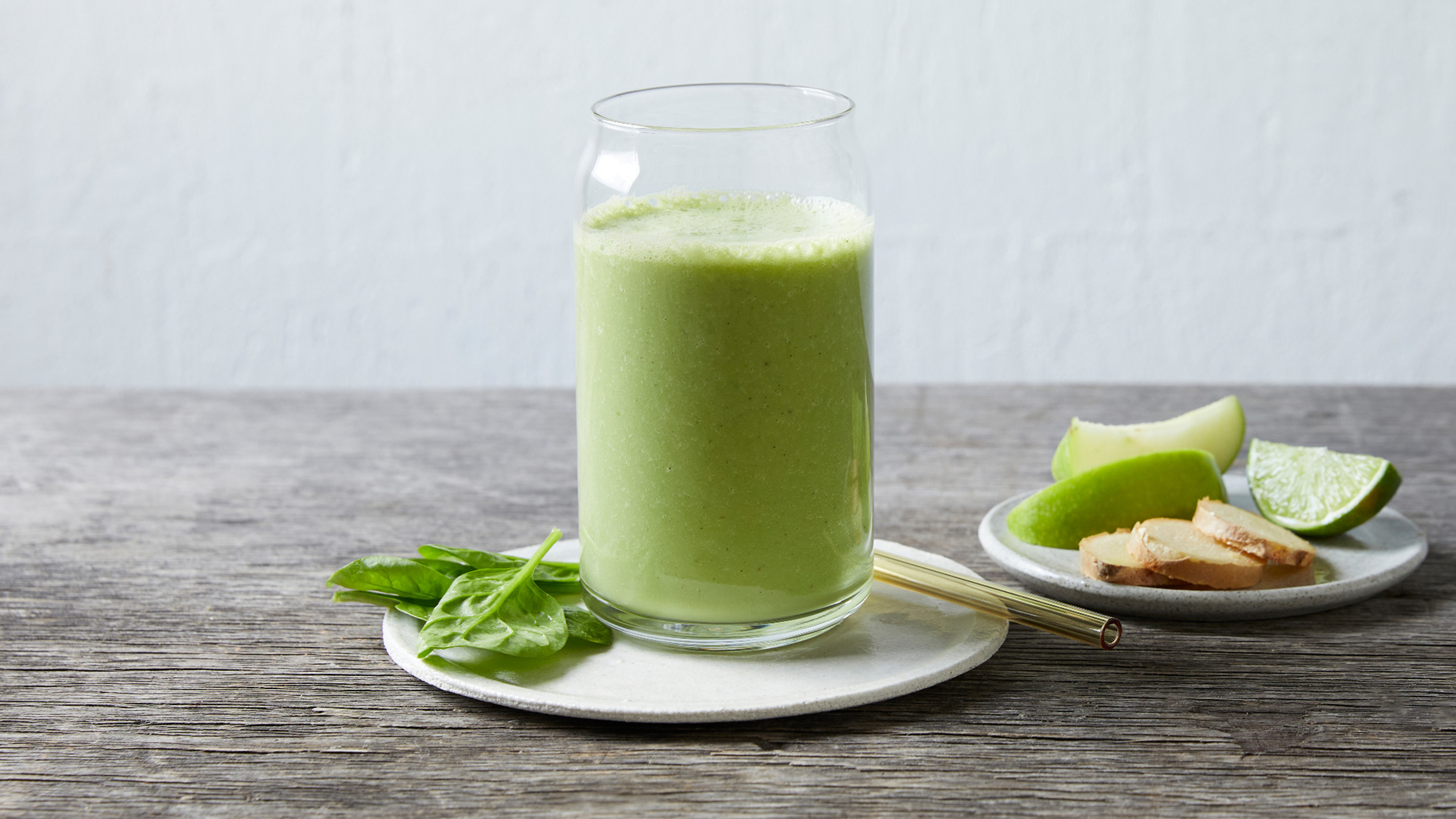A tall glass of green Ginger, apple and banana smoothie sits on small round plate with a metal straw and some basil leaves on the side.