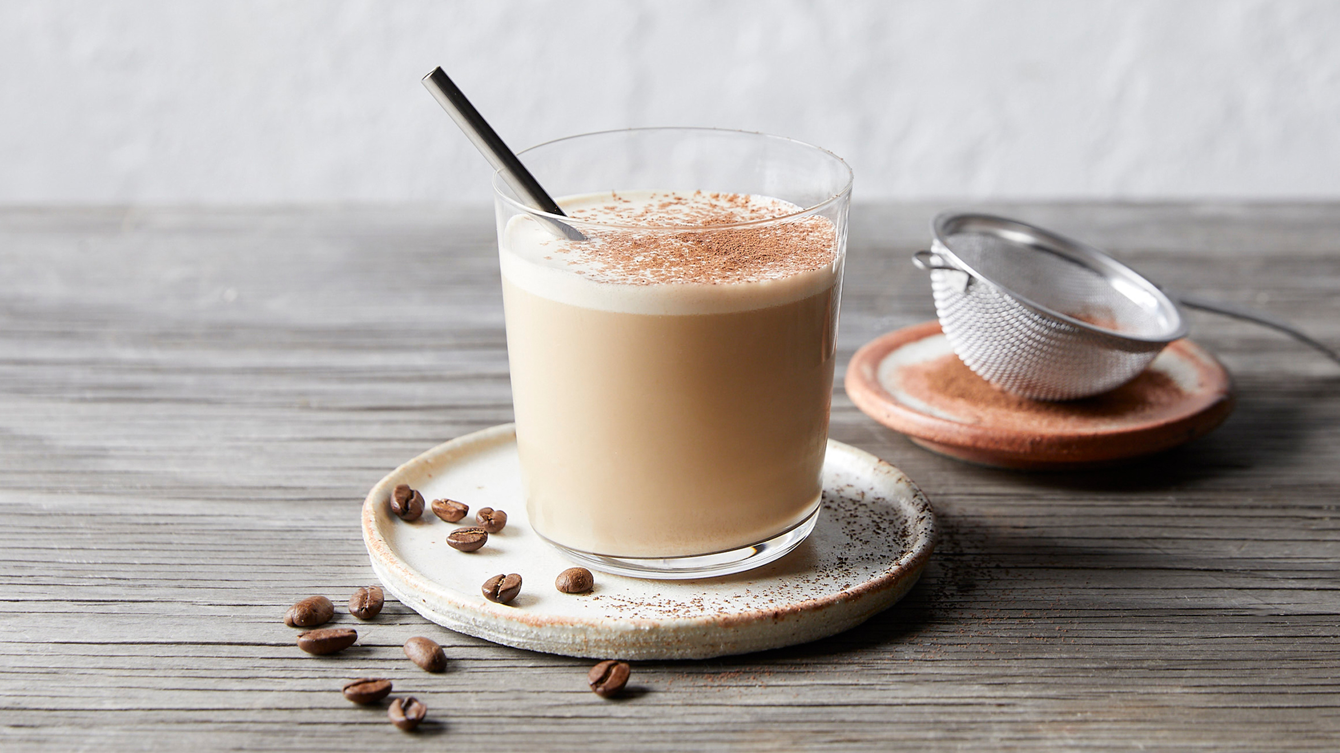 A squat glass of creamy Maple Coffee Smoothie sits on a round white stoneware plate, with coffee beans scattered around
