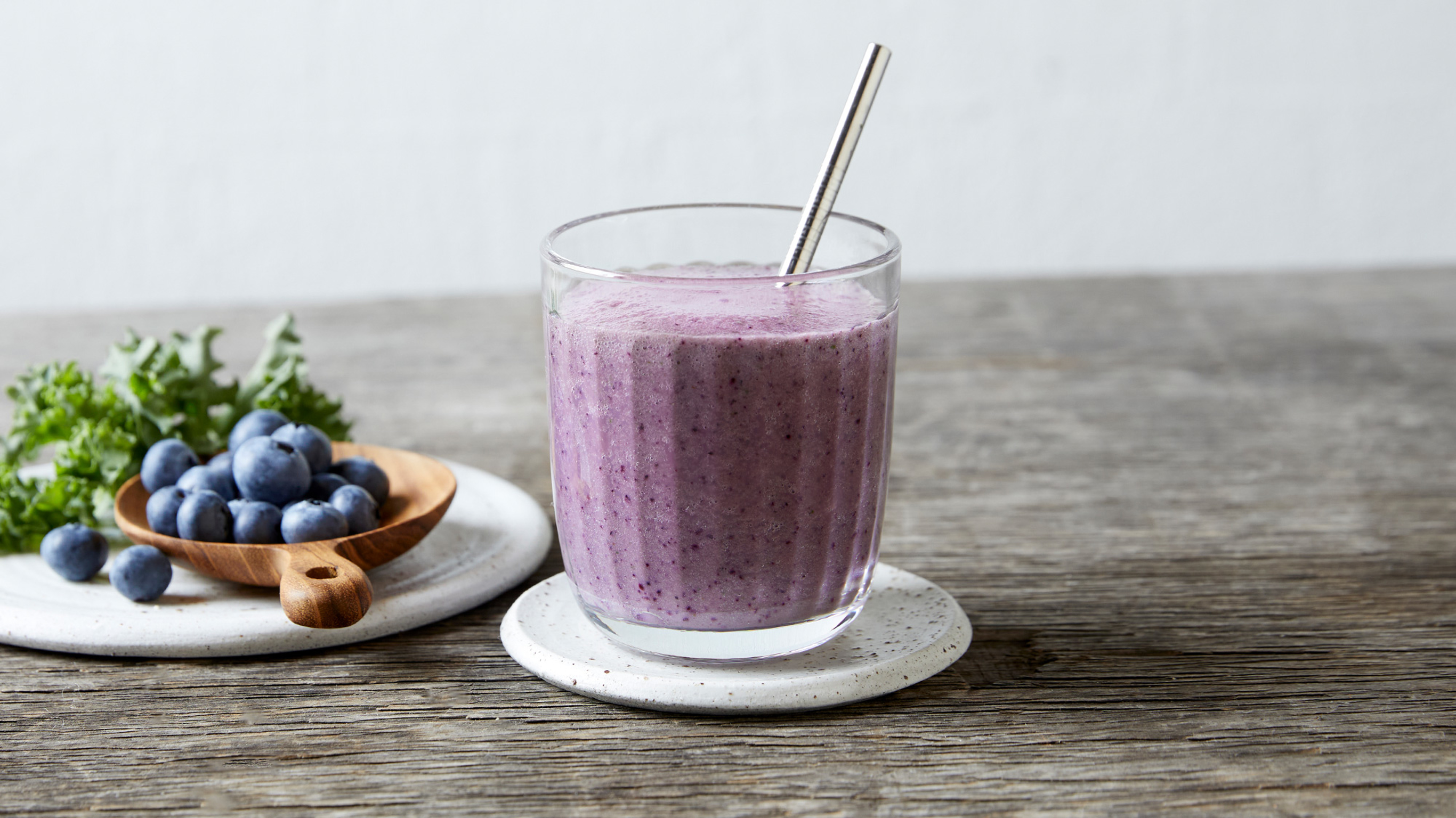A small glass of purple Blueberry and Kale smoothie rests on a small white stoneware plate with some blueberries and kale in the background.