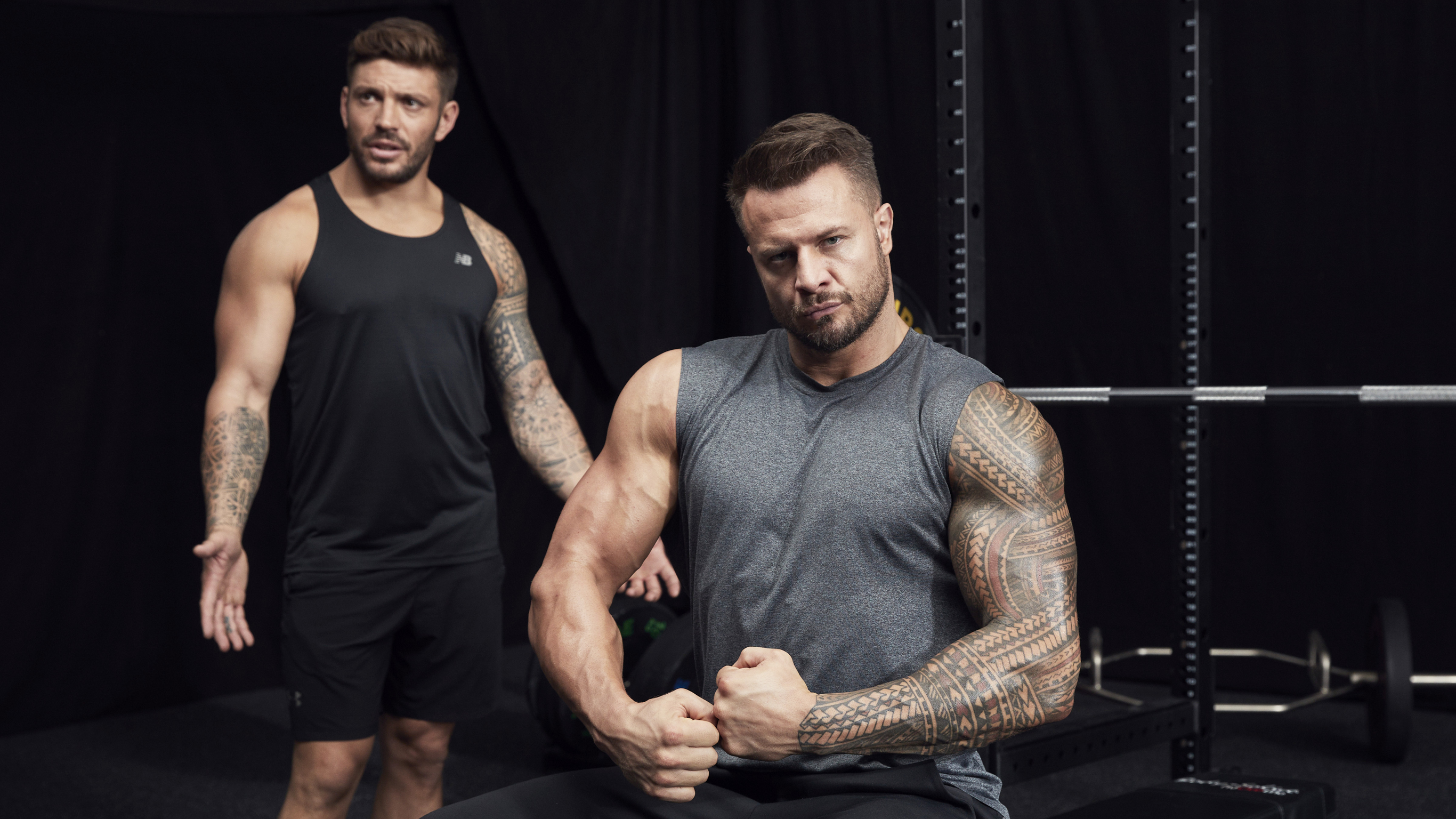 Bobby Holland Hanton poses for the camera, biceps flexed, looking strong, while in the background, Luke Zocchi holds his hands out, querying Bobby's pride in his biceps.