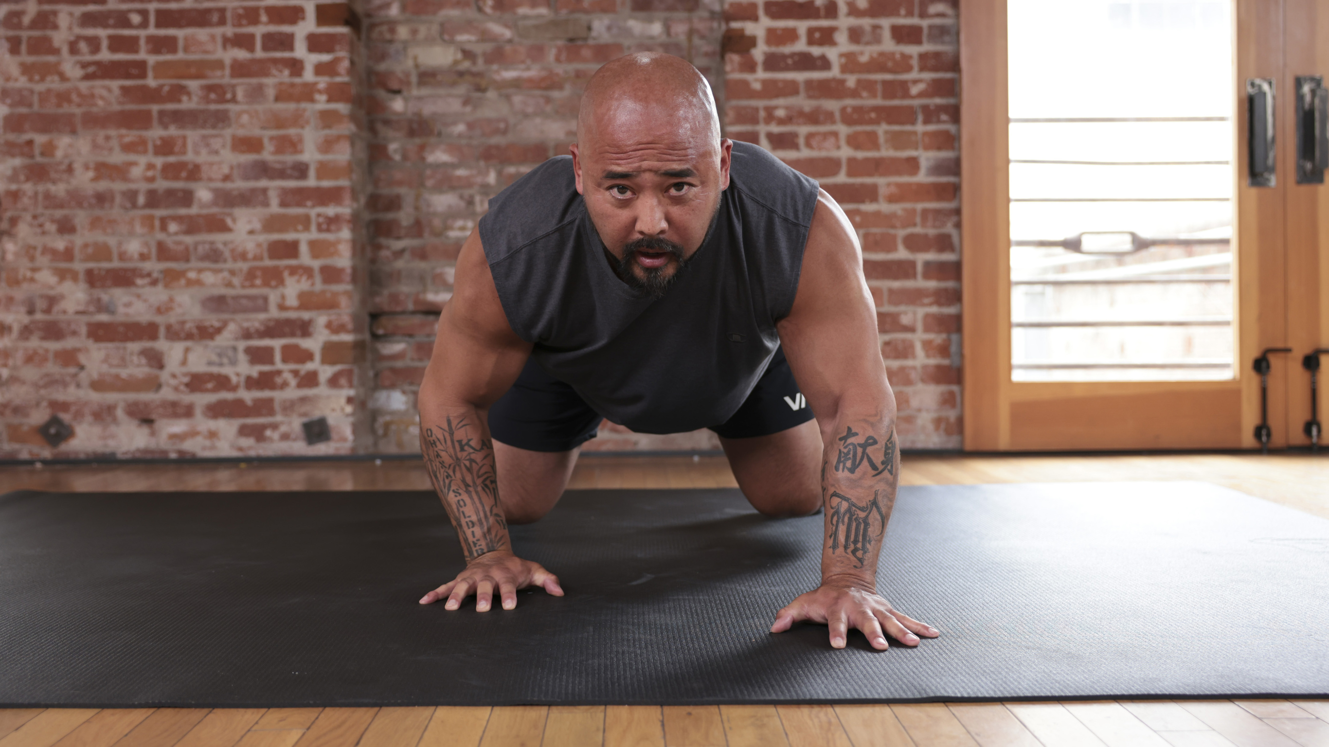 Centr trainer Joseph ‘Da Rulk’ Sakoda, wearing gray and looking directly into the camera with an intense expression, demonstrates a bear crawl exercise.