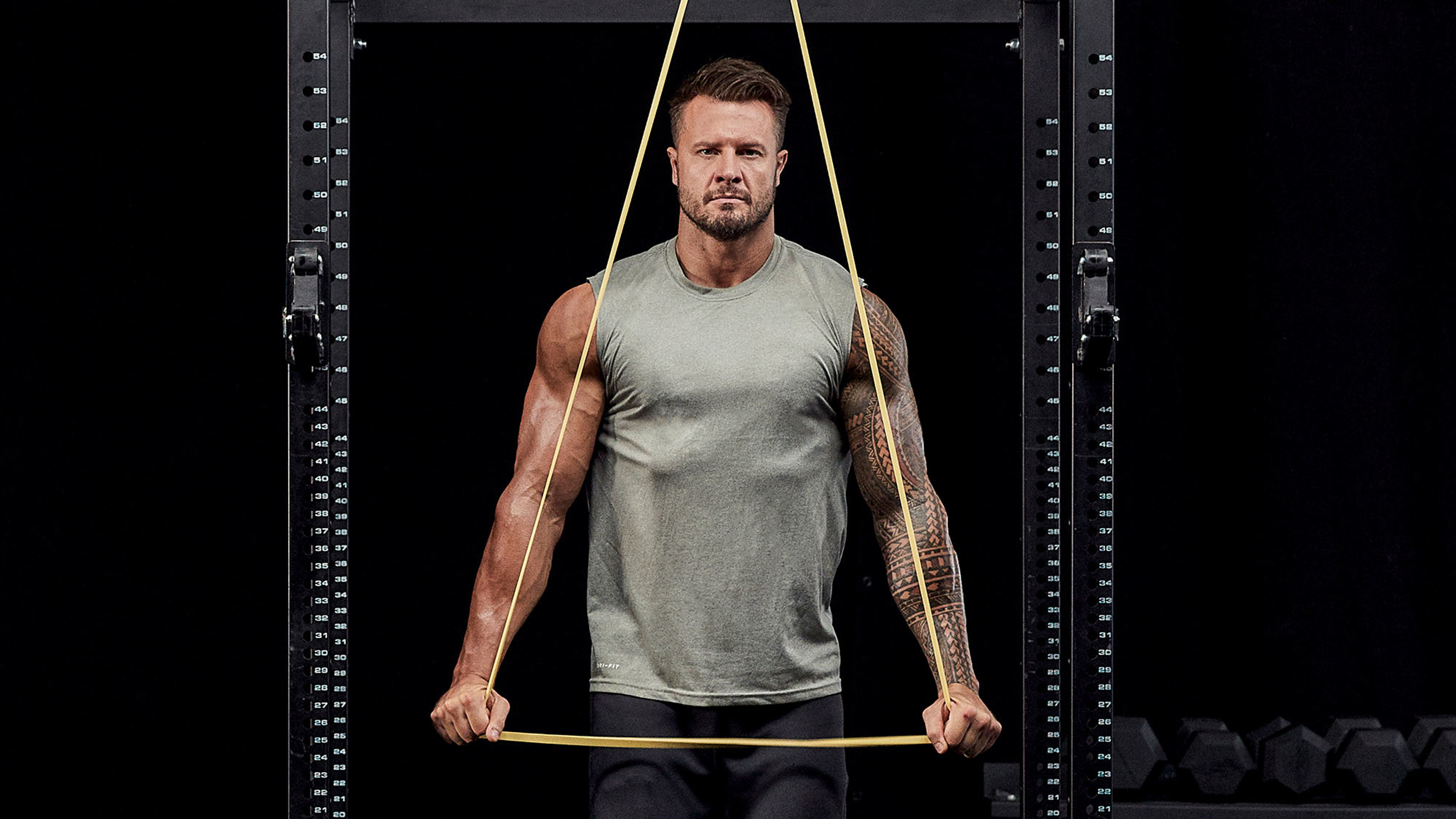Bobby Holland Hanton, wearing a grey tank top, uses a yellow long loop resistance band to perform a lat pulldown exercise from the Centr Power at Home program.