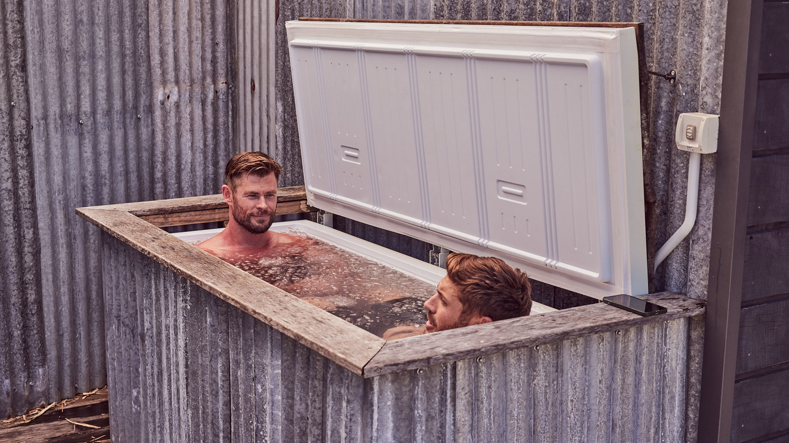Chris Hemsworth and Luke Zocchi sit facing each other, shoulder-deep in an outdoors ice bath.