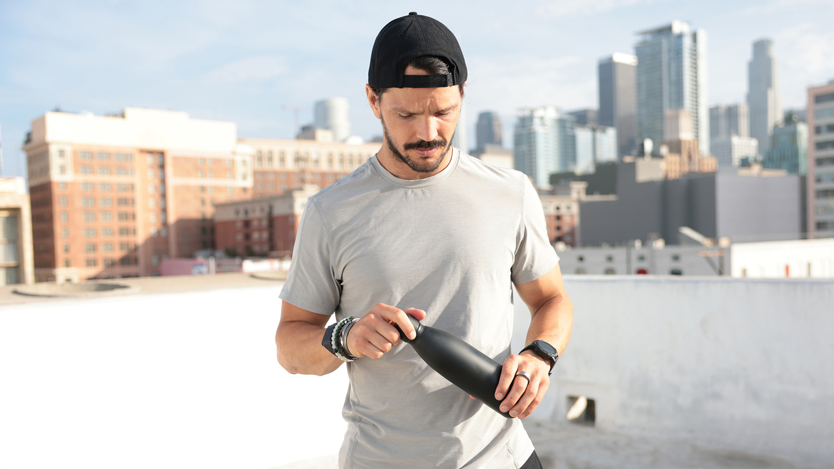Dan Churchill stands on a city roof top, looking down with a black water bottle in his hands, about to unscrew the lid to take a drink of water.