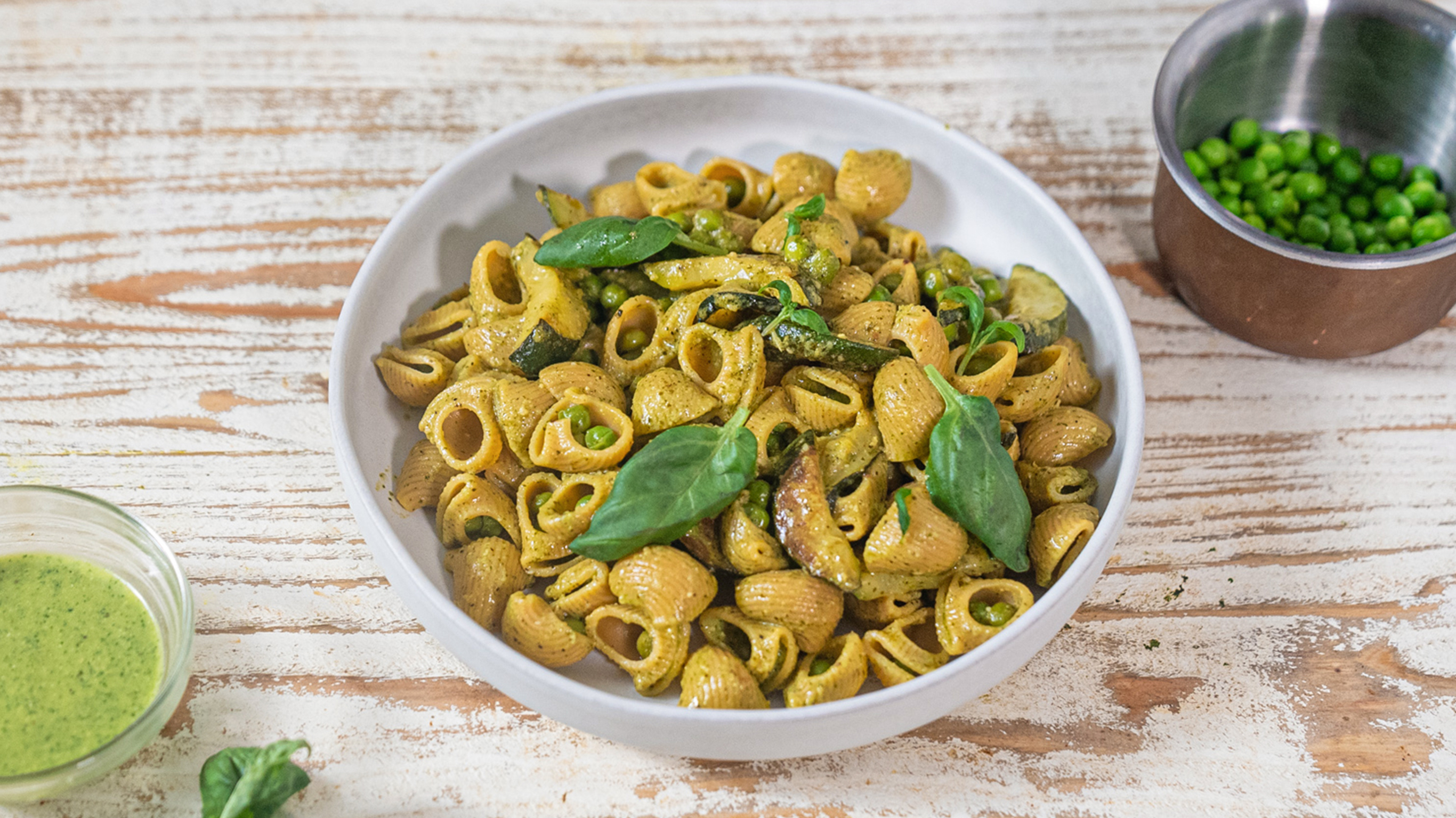 A round plate of Centr's Plant-based Pesto Pasta rests white-washed wooden table, with pesto sausce and small saucepan of peas on the side, ready for eating.