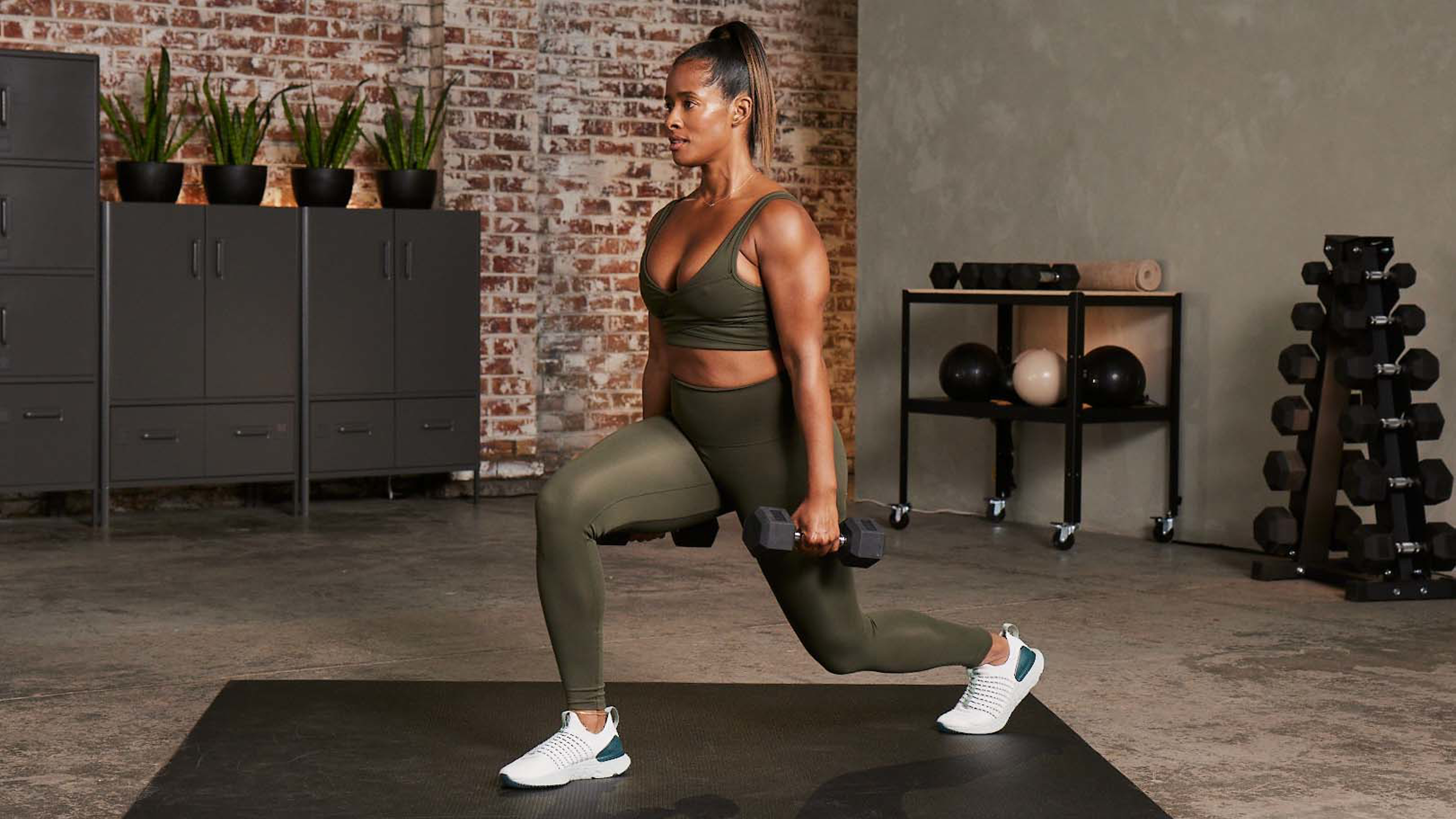 Ingrid Clay performs a forwards lunge with a dumbbell in each hand during a HIIT workout.