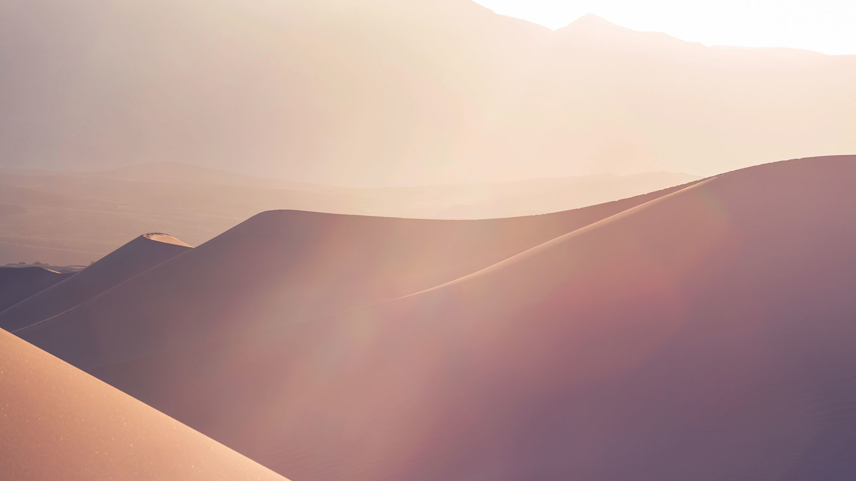 A series of pale red sand dunes are lit by hazy sunlight.