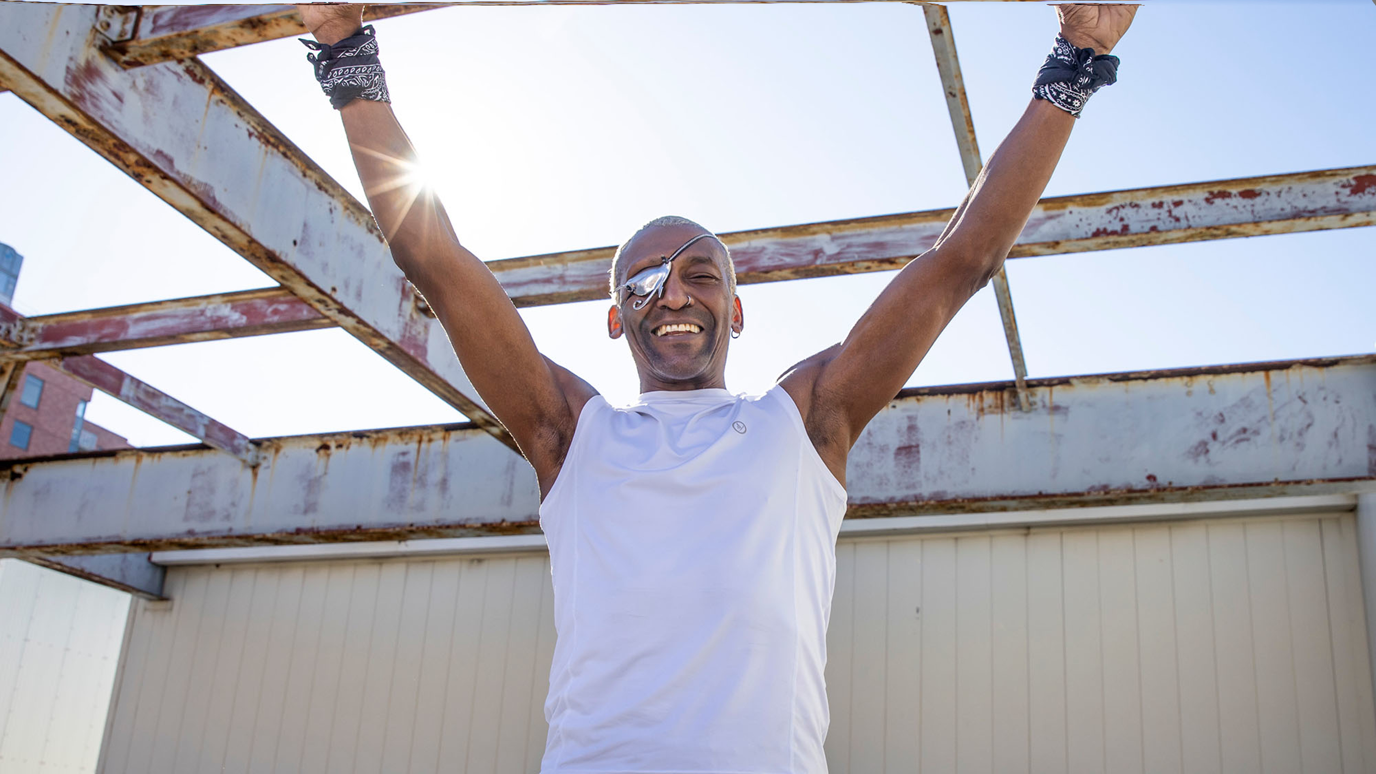 Michael Olajide Jr. wearing a white singlet top raises his arms skyward, standing on an urban rooftop with the sun behind him