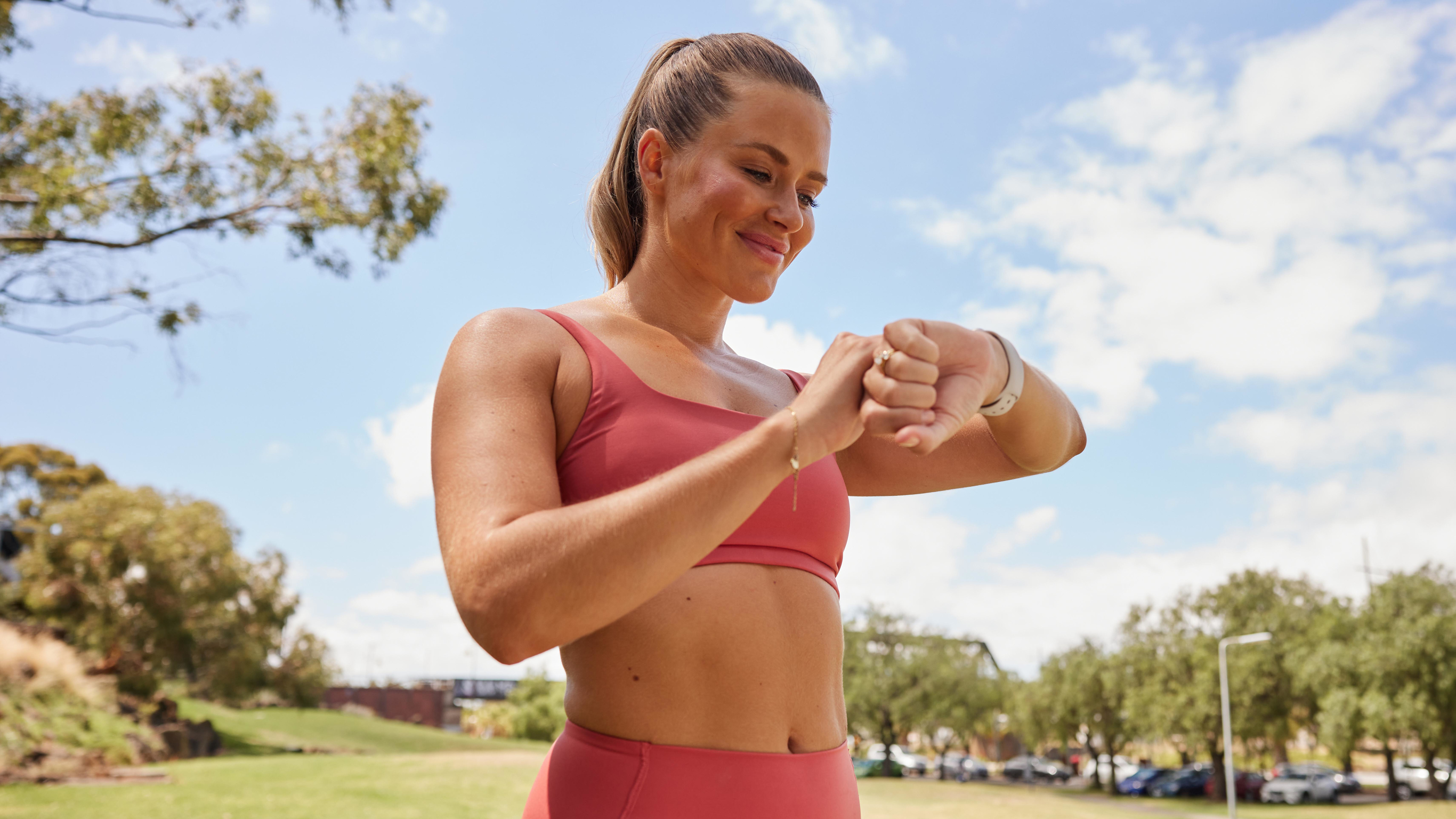 Centr HILIT trainer Alexz Parvi, wearing salmon pink gym wear and standing in a park on a sunny day, looks down at her watch.