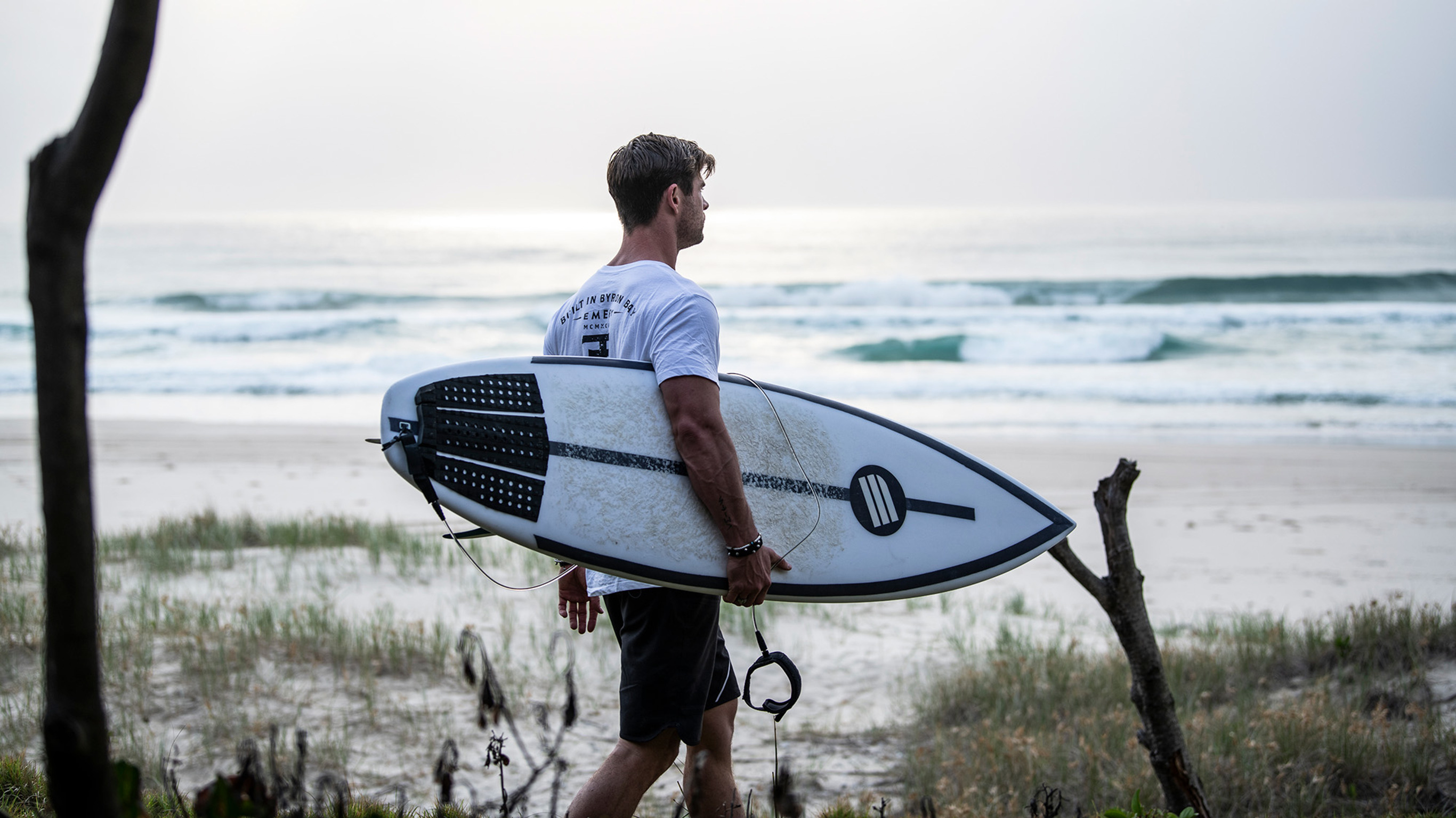 Chris Hemsworth, in a white t-shirt and dark board shorts, carries his surfboard under his right arm as he walks on to the beach at day break.