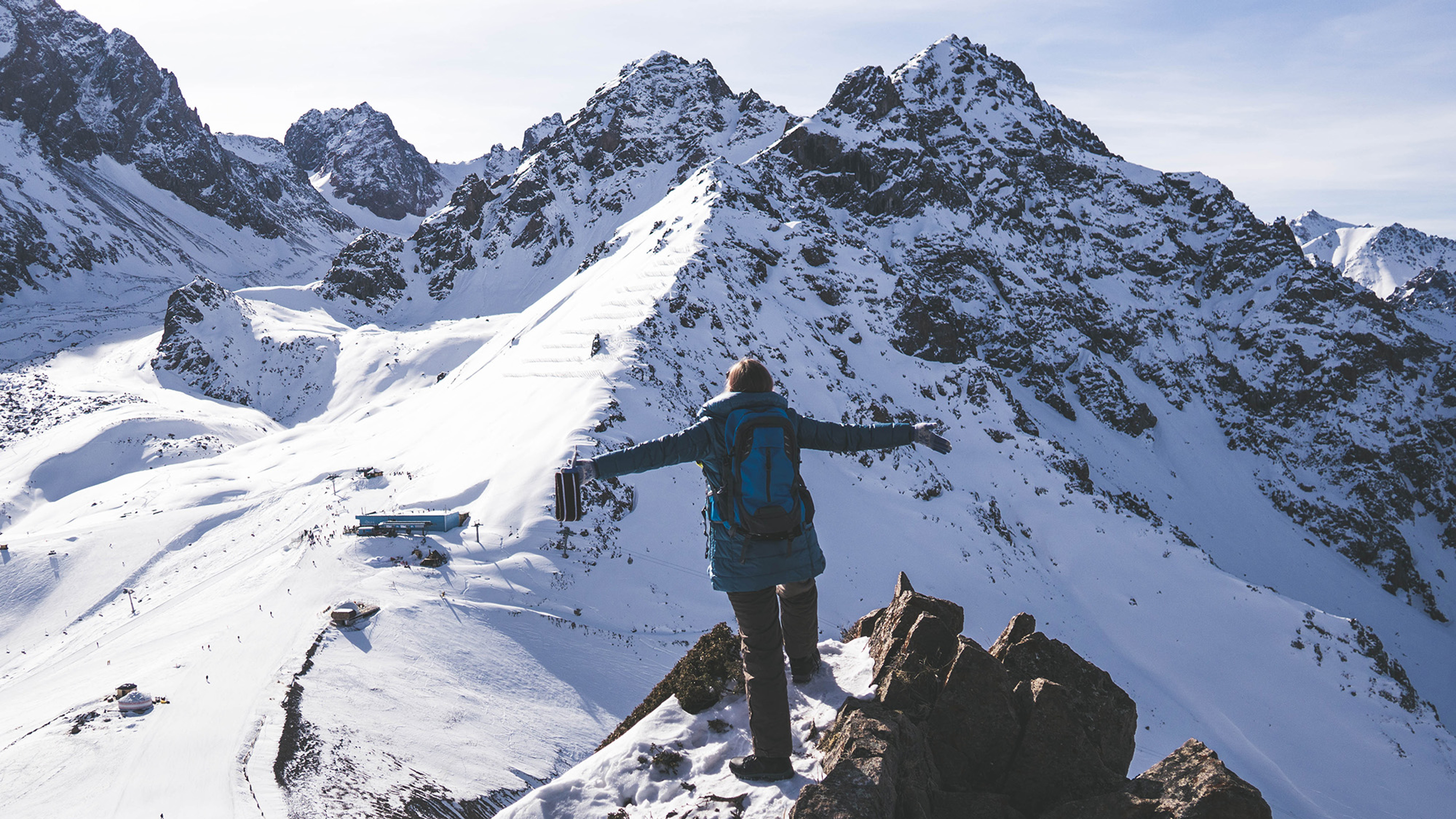 A person stands on a ridge peak, arms outstretched, facing a taller mountain peak ahead of them.