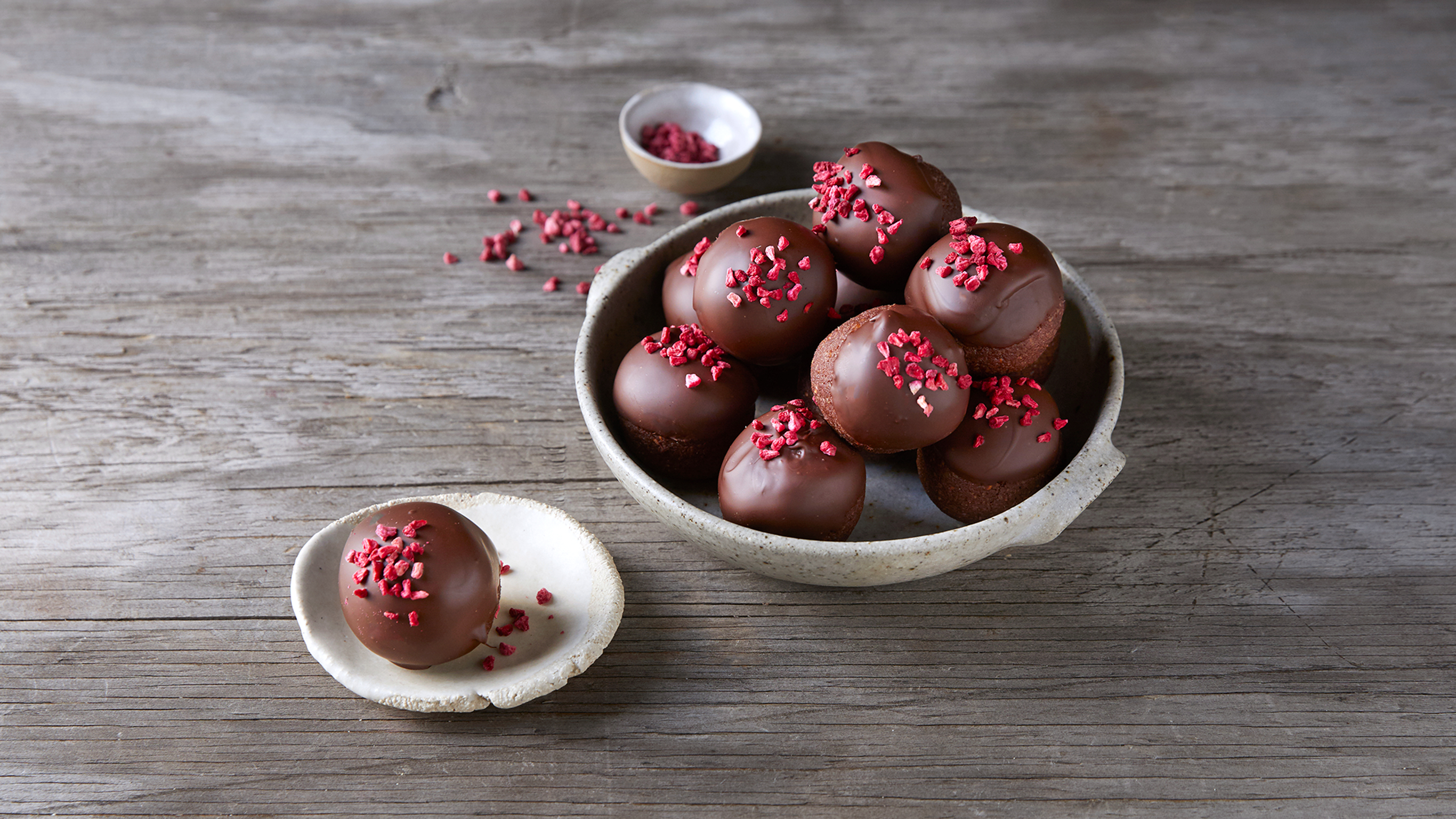 A white bowl of chocolate truffles from the Centr meal plan, decorated with raspberries, rests on a wooden table.