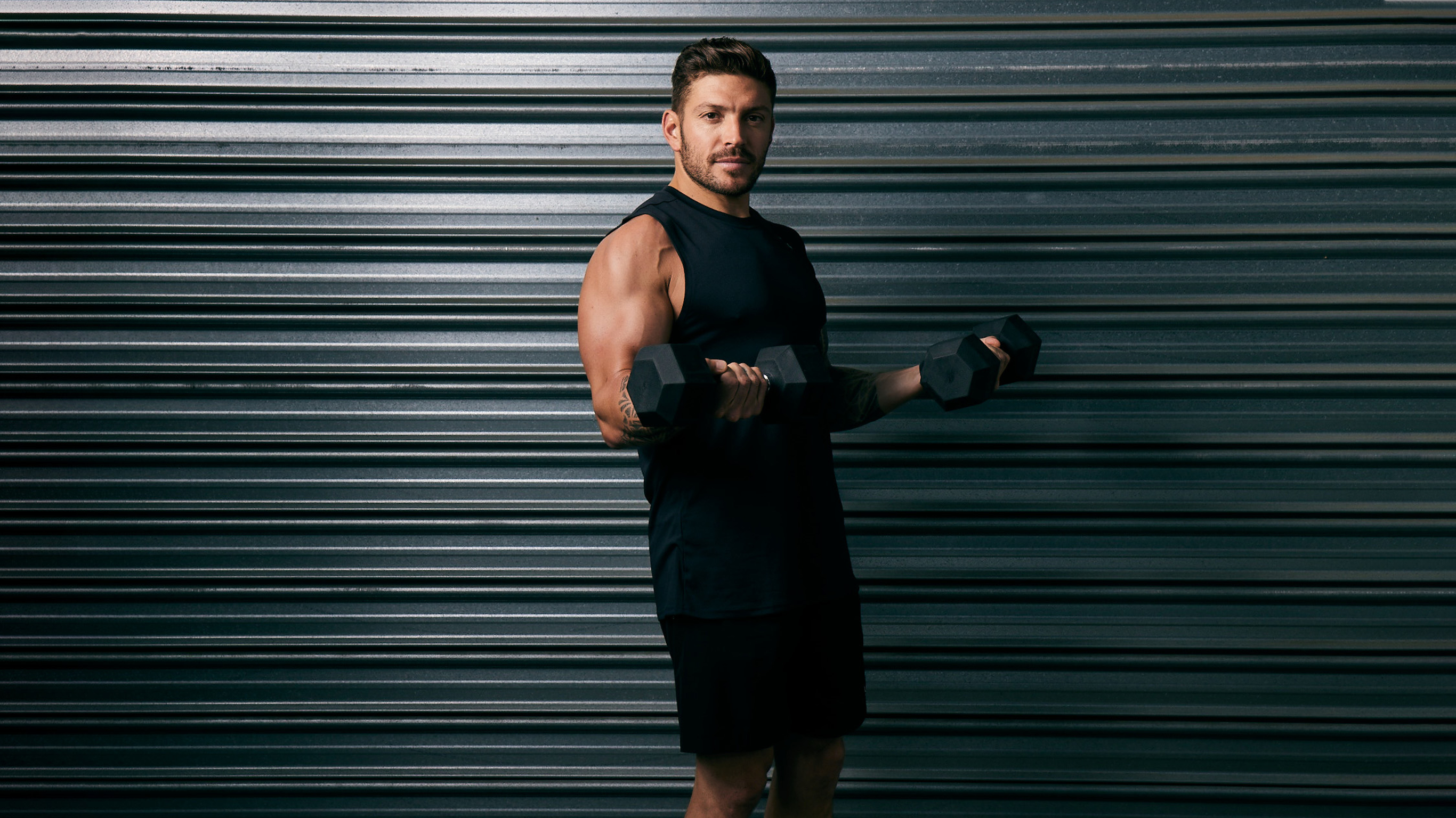 Luke Zocchi stands in front of a dark metal grey roller door, holding a dumbbell in each hand in a bicep curl, looking at the camera.