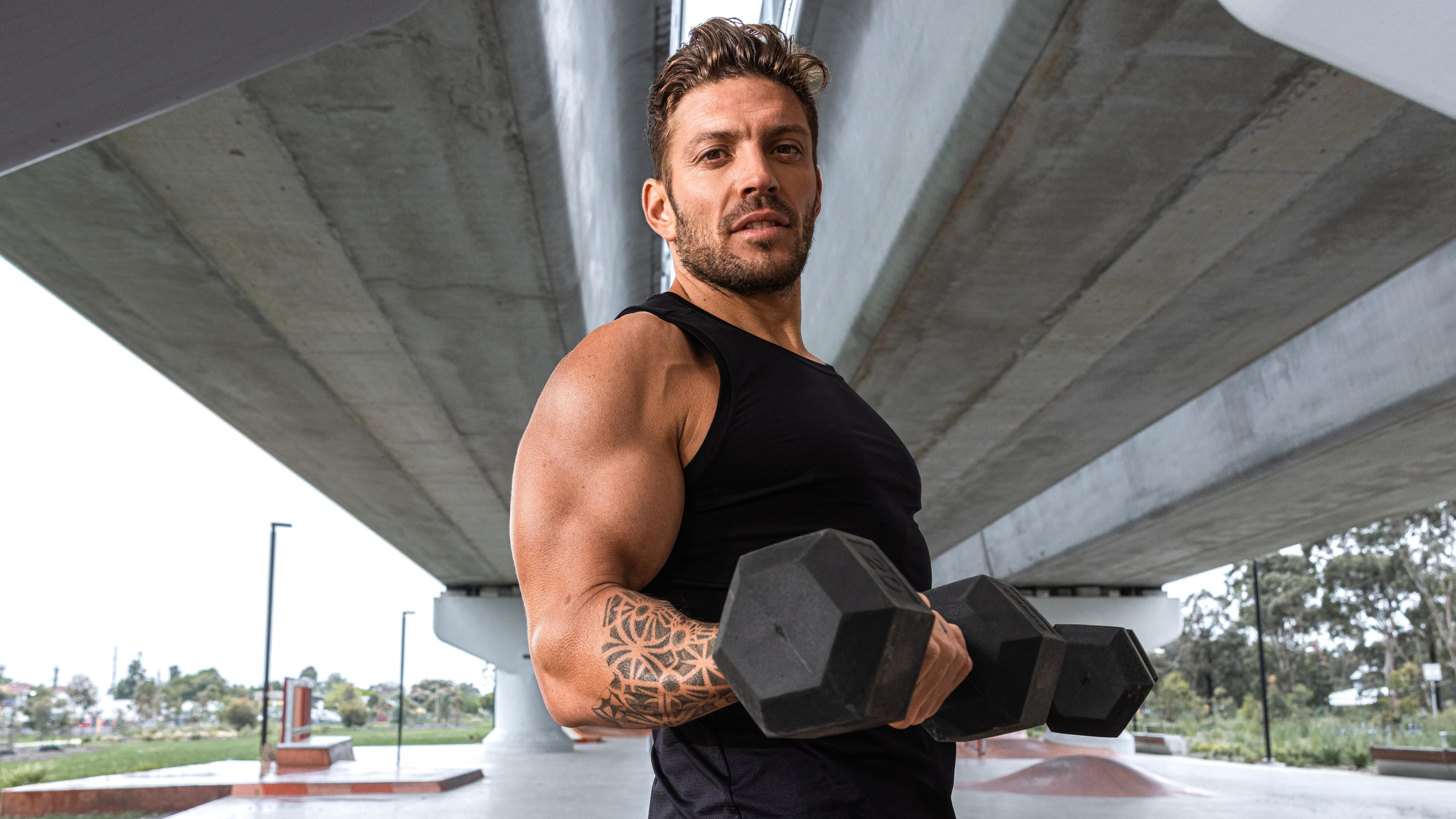 Centr trainer Luke Zocchi, standing under a concrete bridge and wearing a muscle shirt, holds two dumbbells in a bicep curl, looking down at the camera.