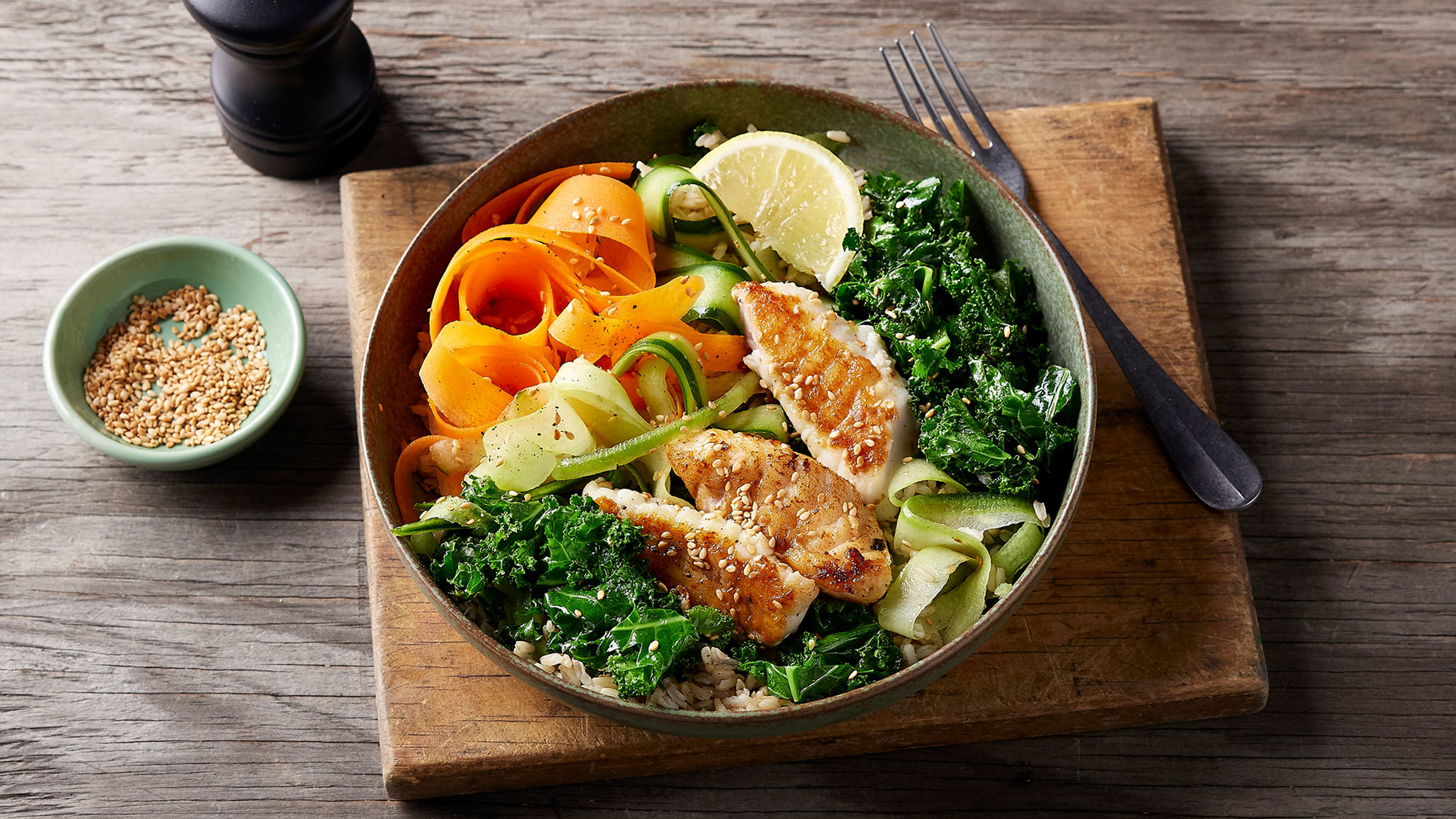 A bowl of grilled fish and fresh vegetables sits on a table.