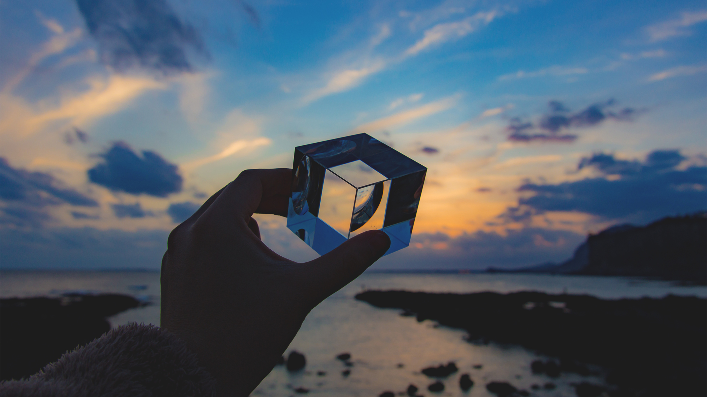 An image of a hand holding a clear cube up towards the setting sun at a rocky beach.