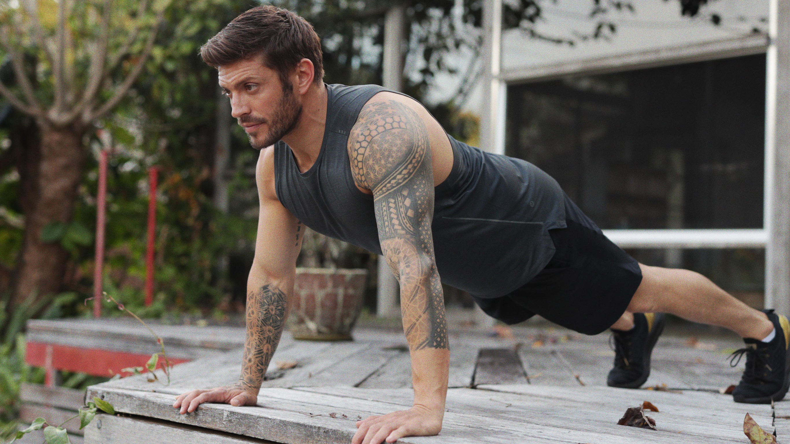 Centr trainer Luke Zocchi performs a full plank outside, with his hands on the edge of a wooden verandah.