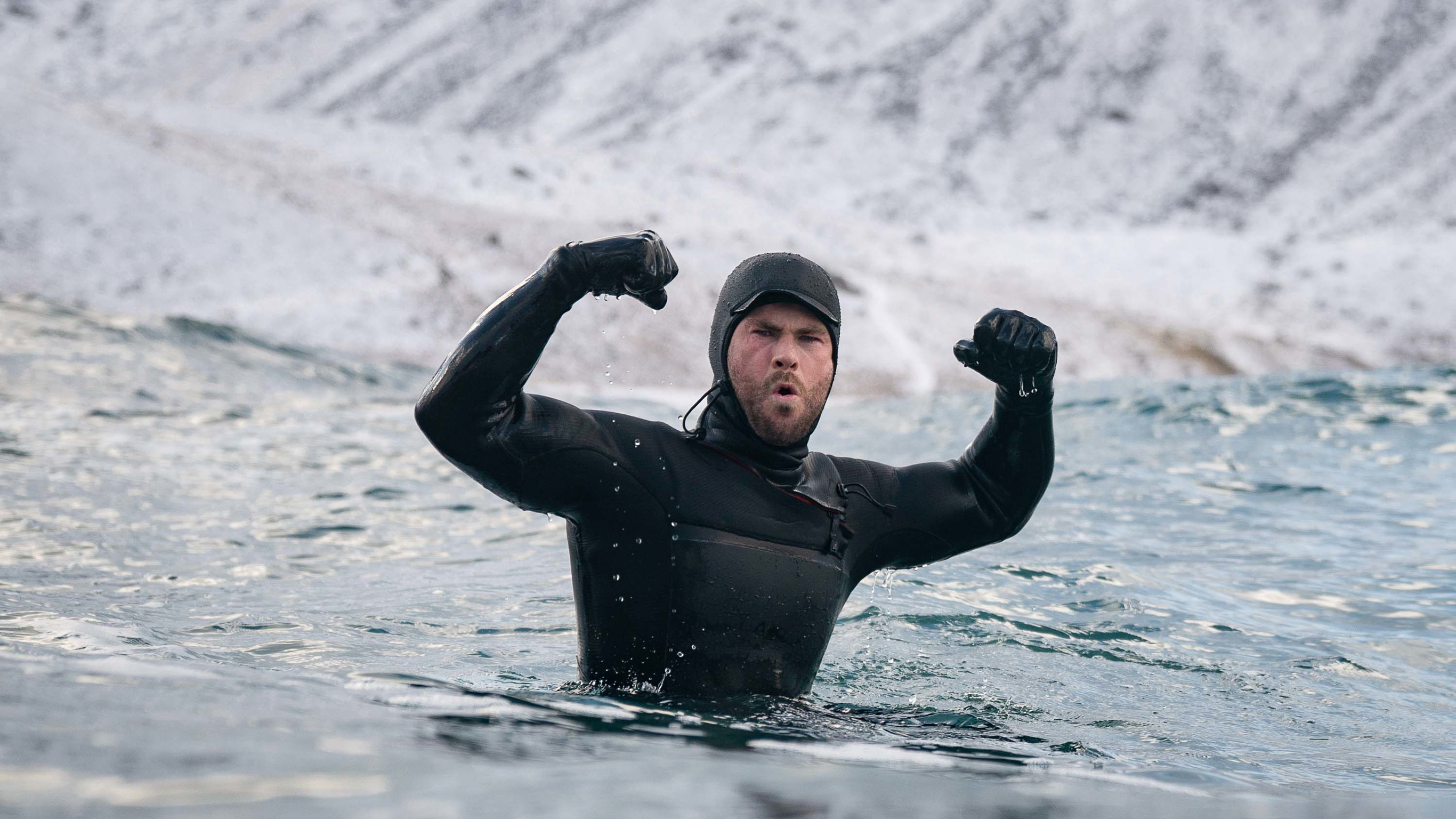 Chris Hemsworth, wearing a full-body wetsuit, stands arms raised and flexed in icy, turbulent ocean waves.