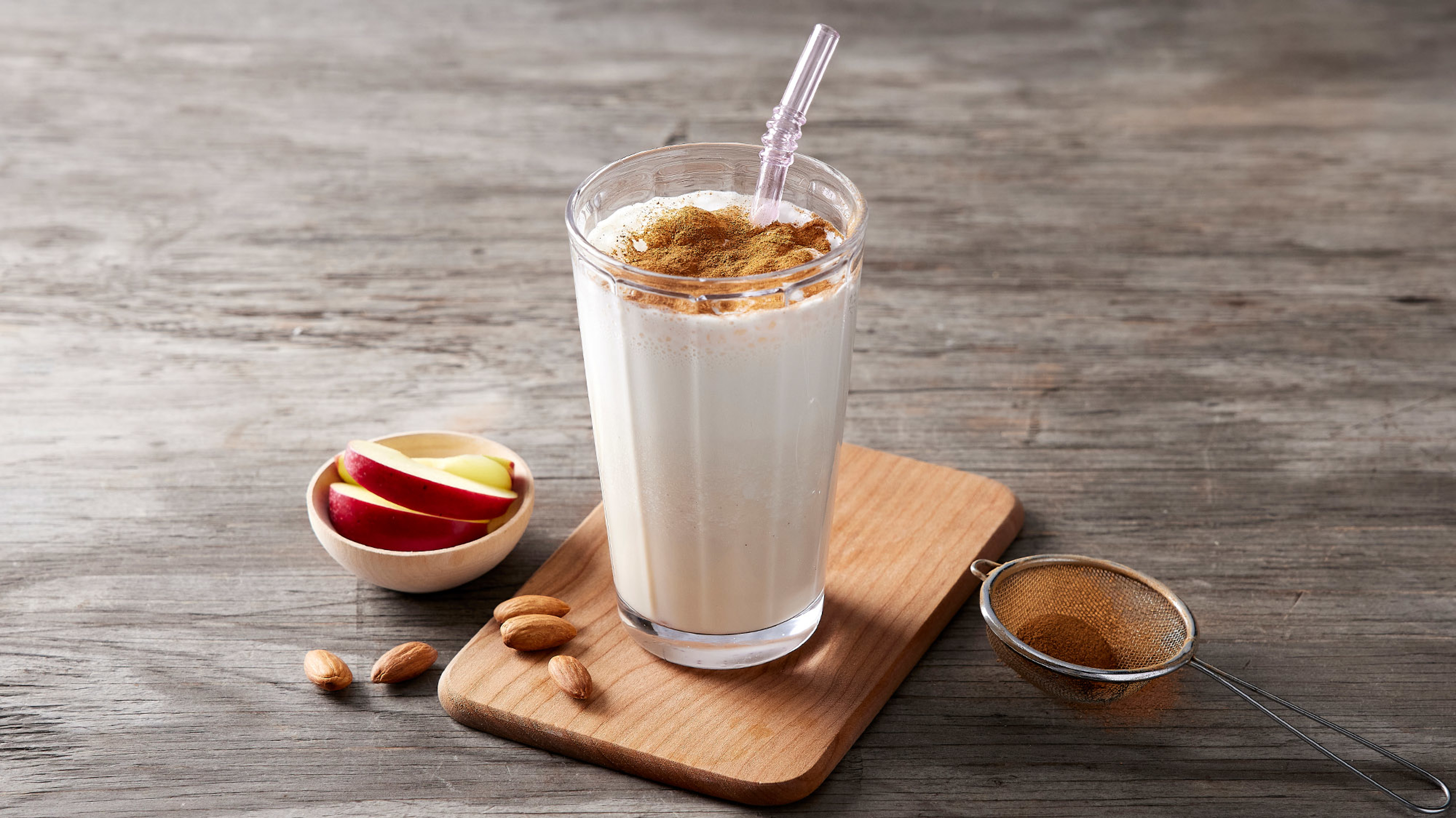 A glass of milky smoothie dusted with cocoa and with a glass straw sits on a wooden tray beside a small bowl of sliced apple.