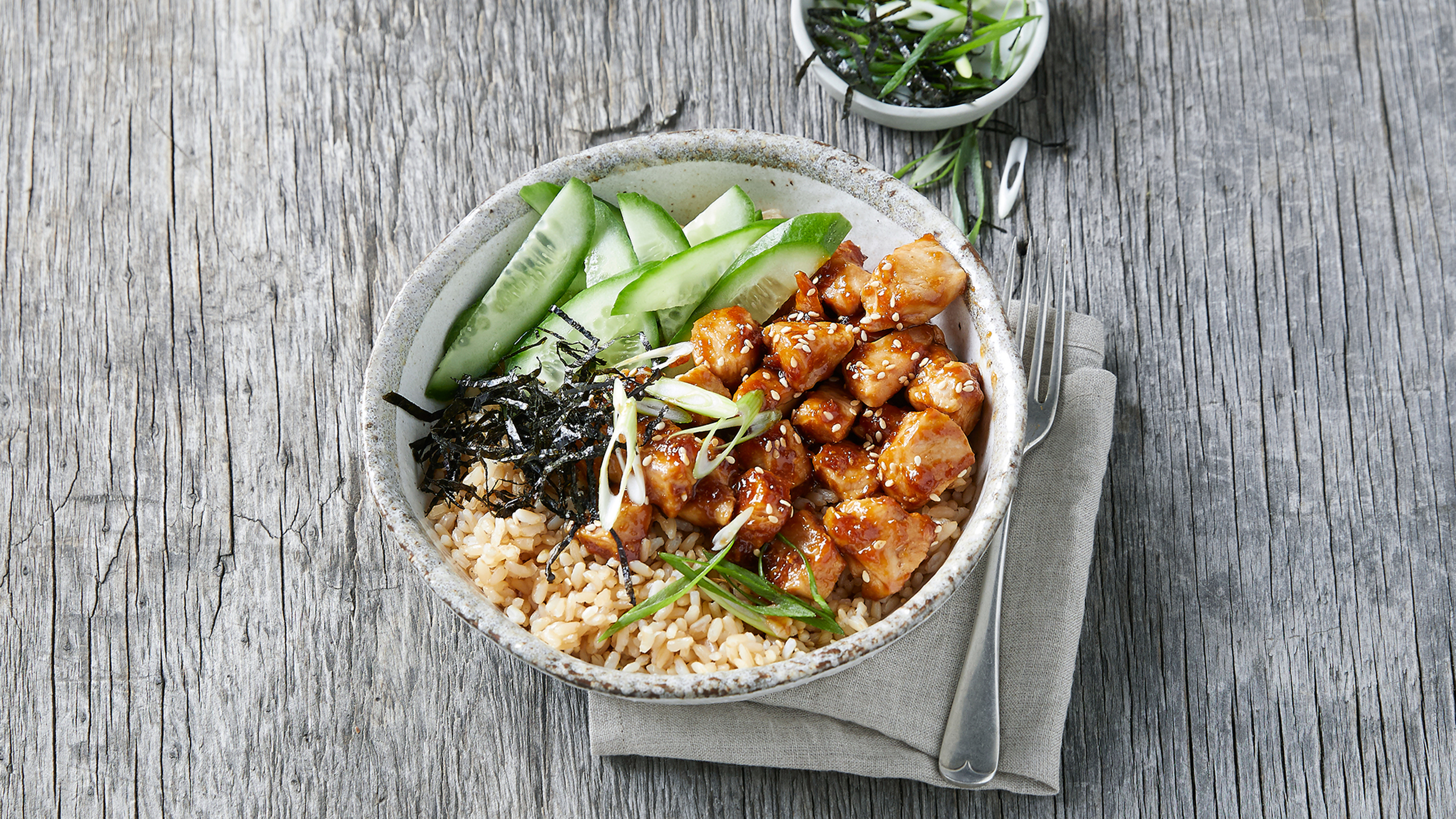 A bowl of teriyaki chicken, rice, cucumber and shredded nori sits on a greyed wooden table.