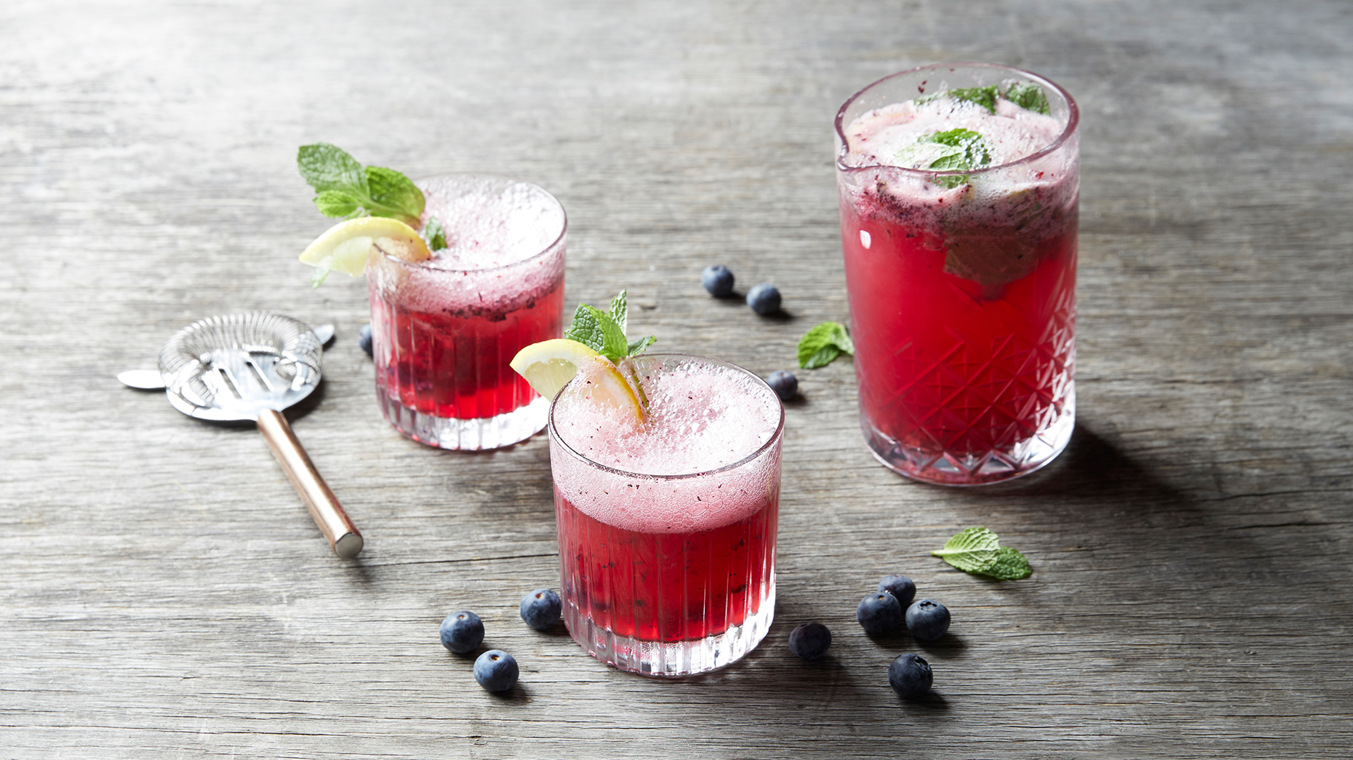 3 glasses of Centr's blueberry lemon spritzer., garnished with lemon slices and mint, sit on a rustic grey wooden table.