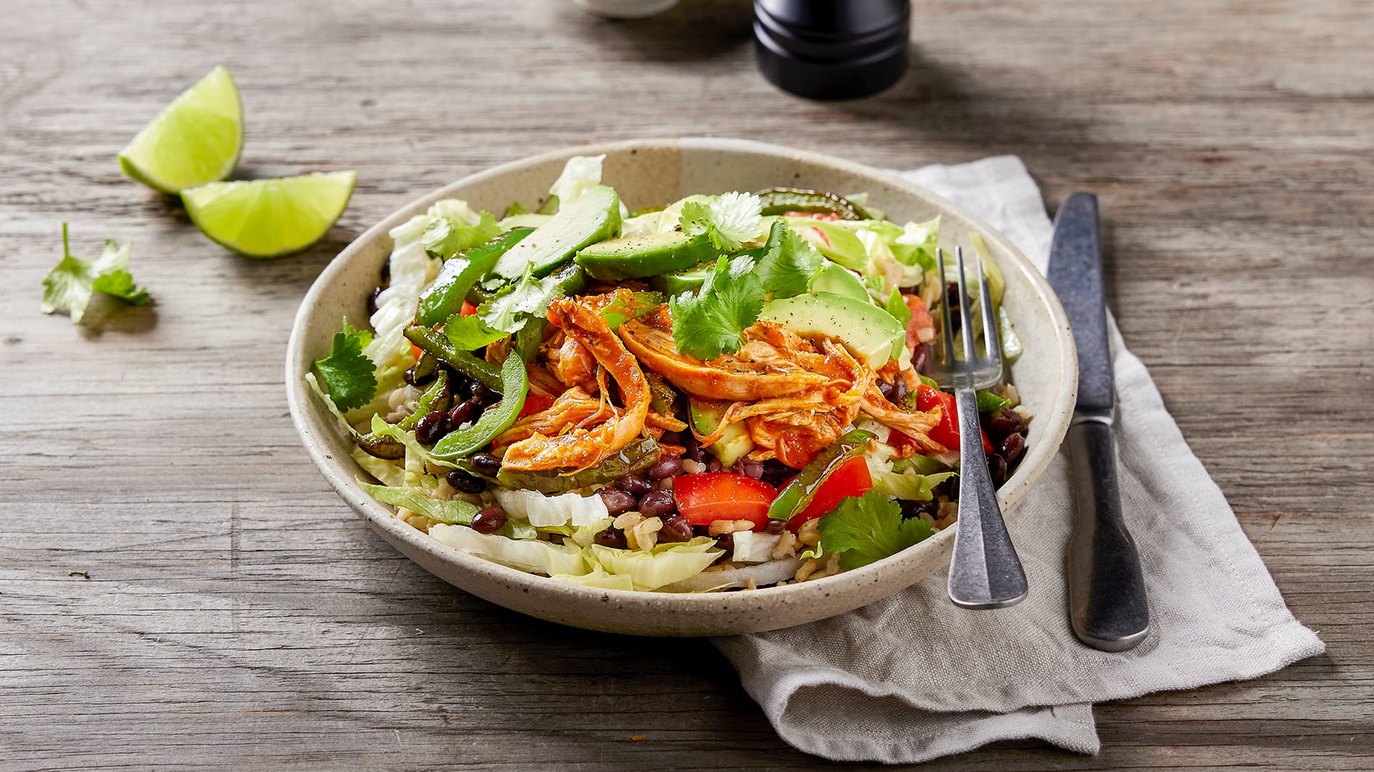 A bowl of shredded salsa chicken burrito sits on a wooden table with a linen napkin, with a knife and fork on the side.