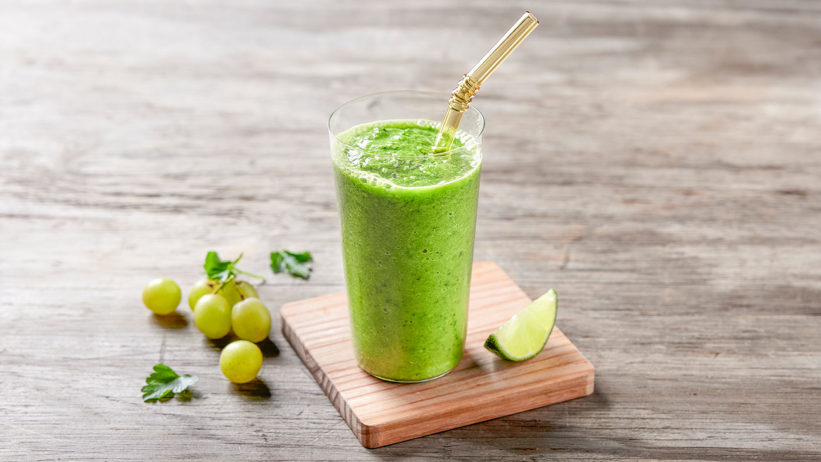 A glass of Zesty Green Smoothie sits on a small wooden board with a quarter of lime on the side.
