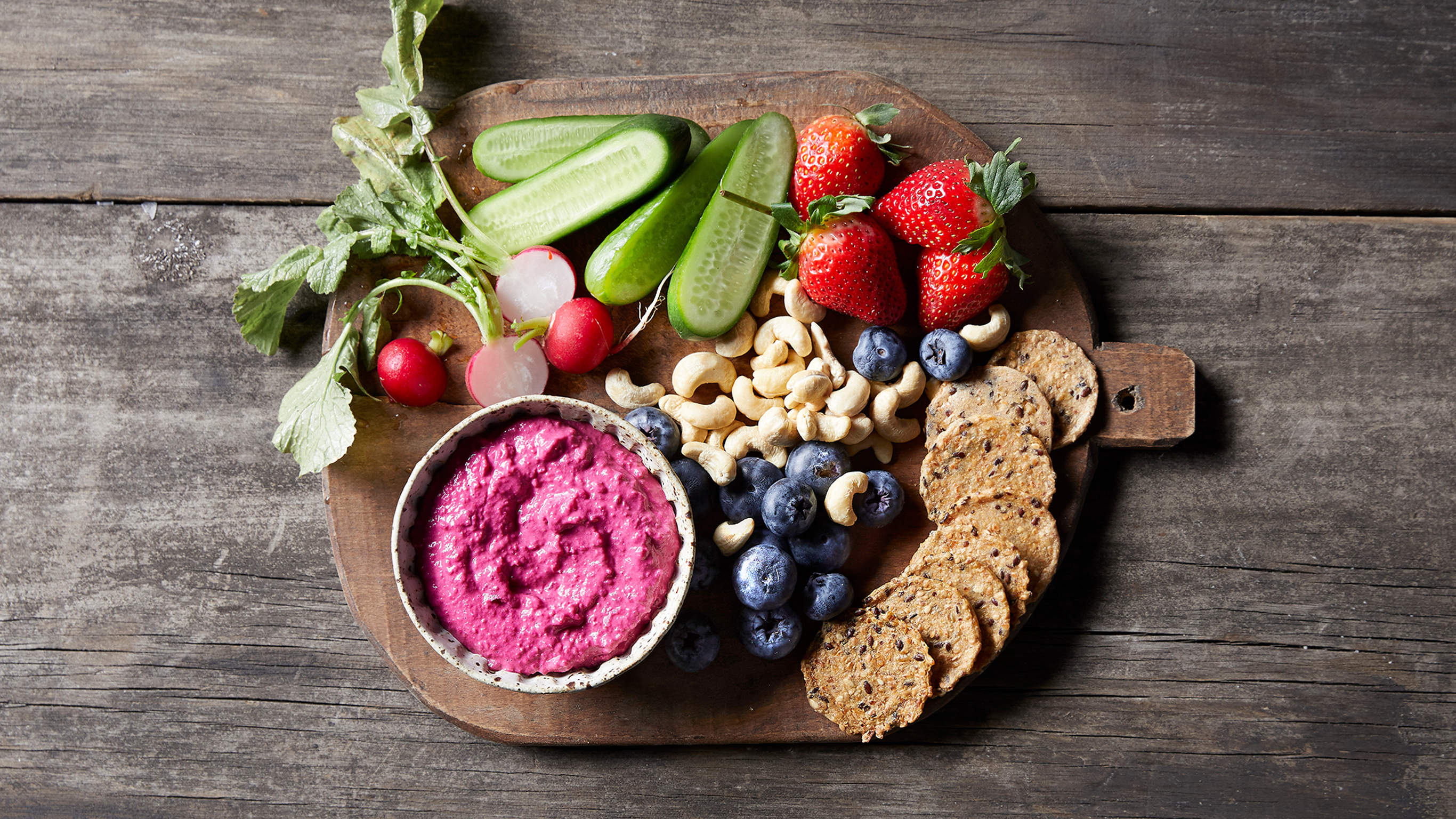 A range of plant-based dip, vegetables, nuts, berries and crackers sit on a wooden cutting board on a wooden table.