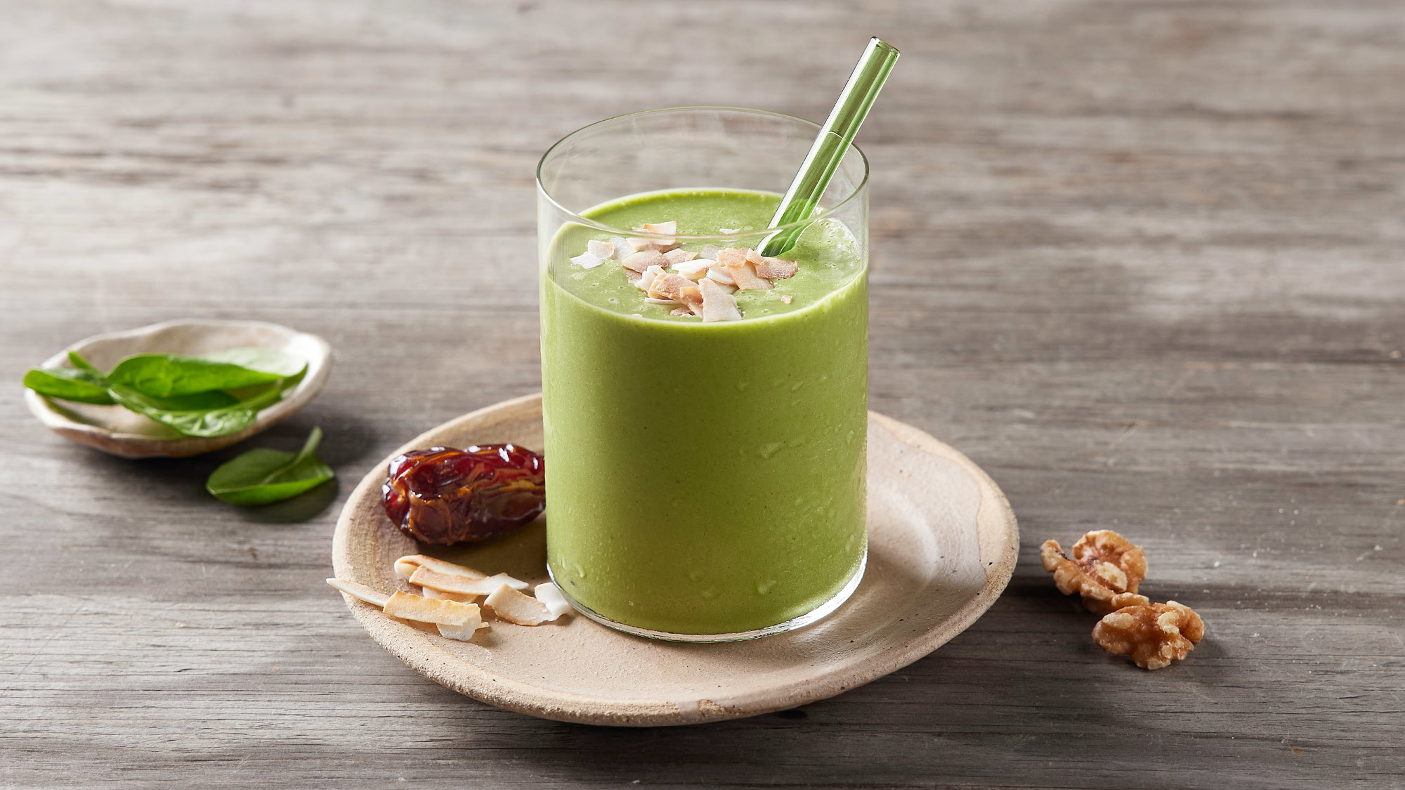 A glass of banana and walnut green smoothie sits on a cream plate with some flaked almonds and a date on the side.