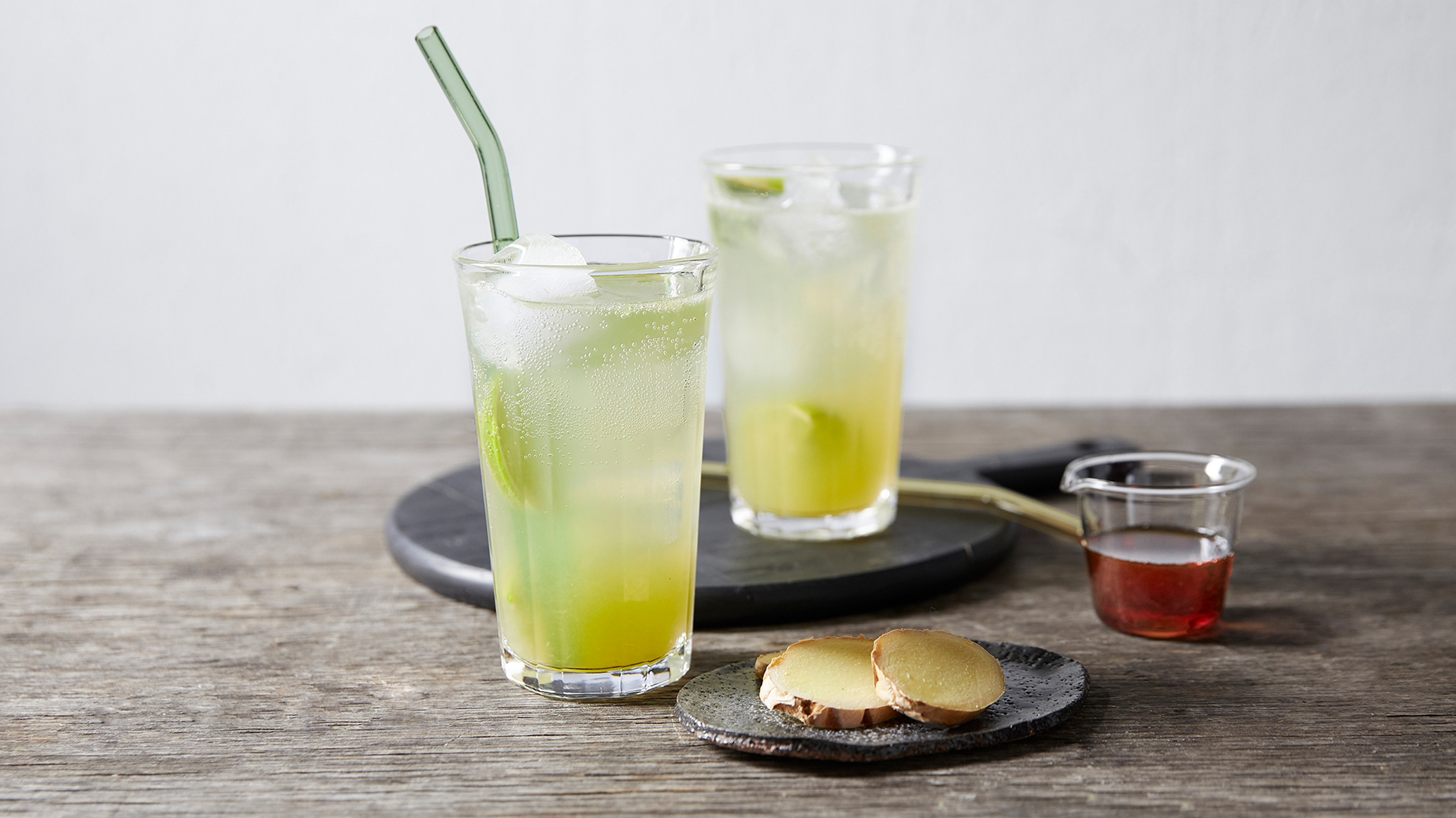 Two tall glasses of pale lime coloured ginger mule mocktail sit on a table with a side dish of ginger slices and a small bowl of maple syrup.