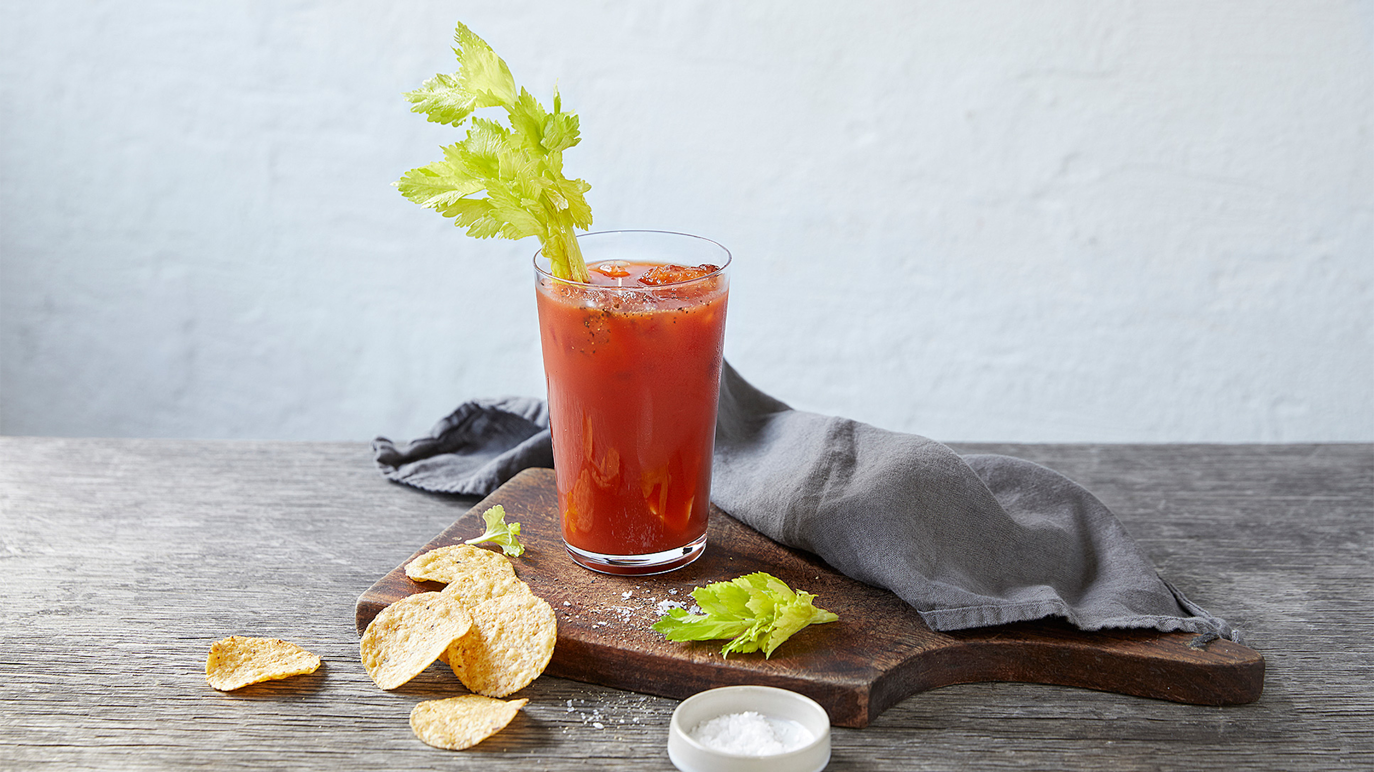 A tall glass of Morning Recovery sits on a wooden board, with a leafy stick of celery in the glass, and some corn chips on the side.