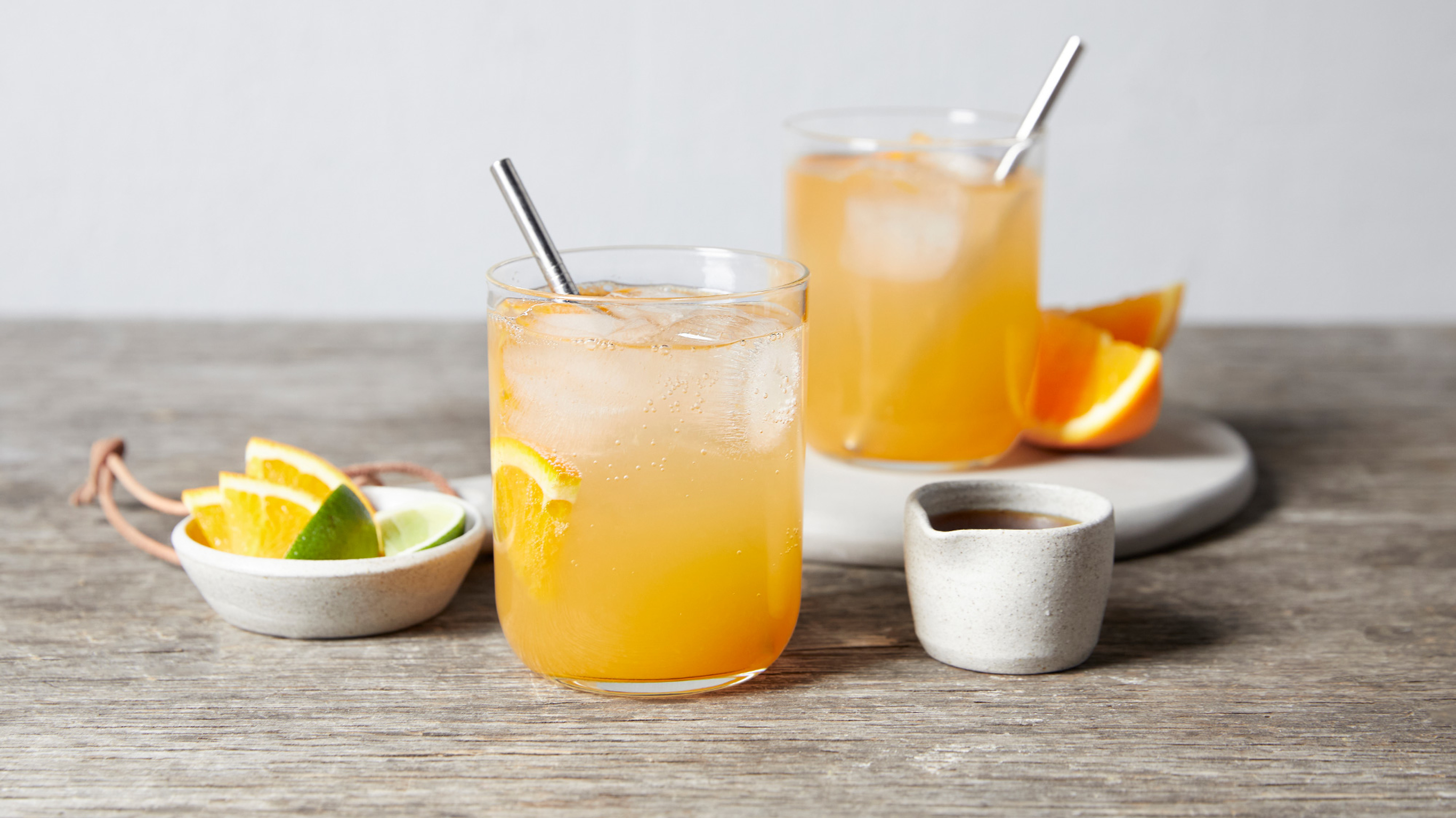2 glasses of Paloma Mocktail sit on a wooden table, with citrus quarters in a side dish and a small bowl of maple syrup on the side.